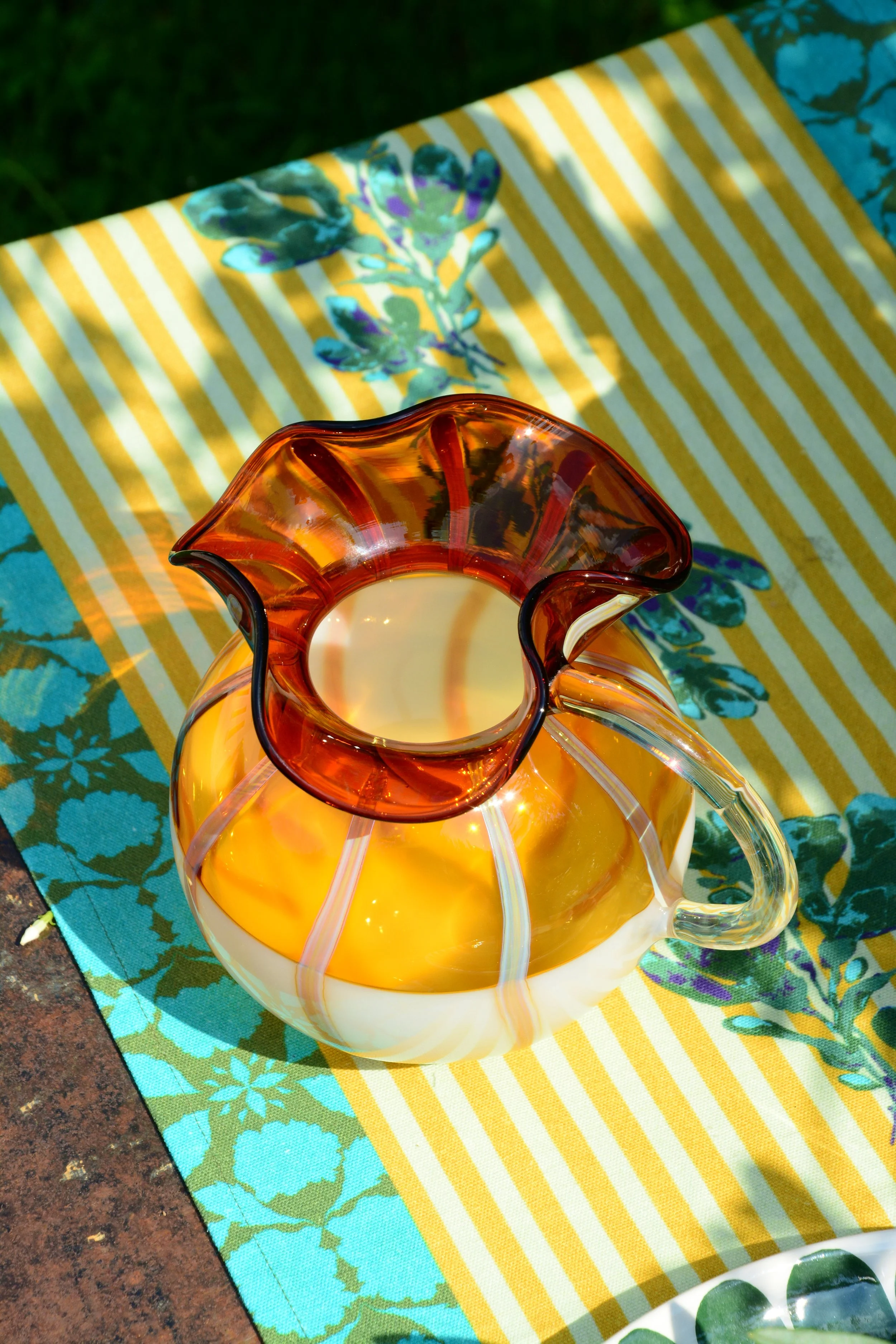 A glass vase with an orange and white swirl design, containing a lit candle, sits on a colorful tablecloth with yellow and white stripes and floral patterns, outdoors on a brick surface.