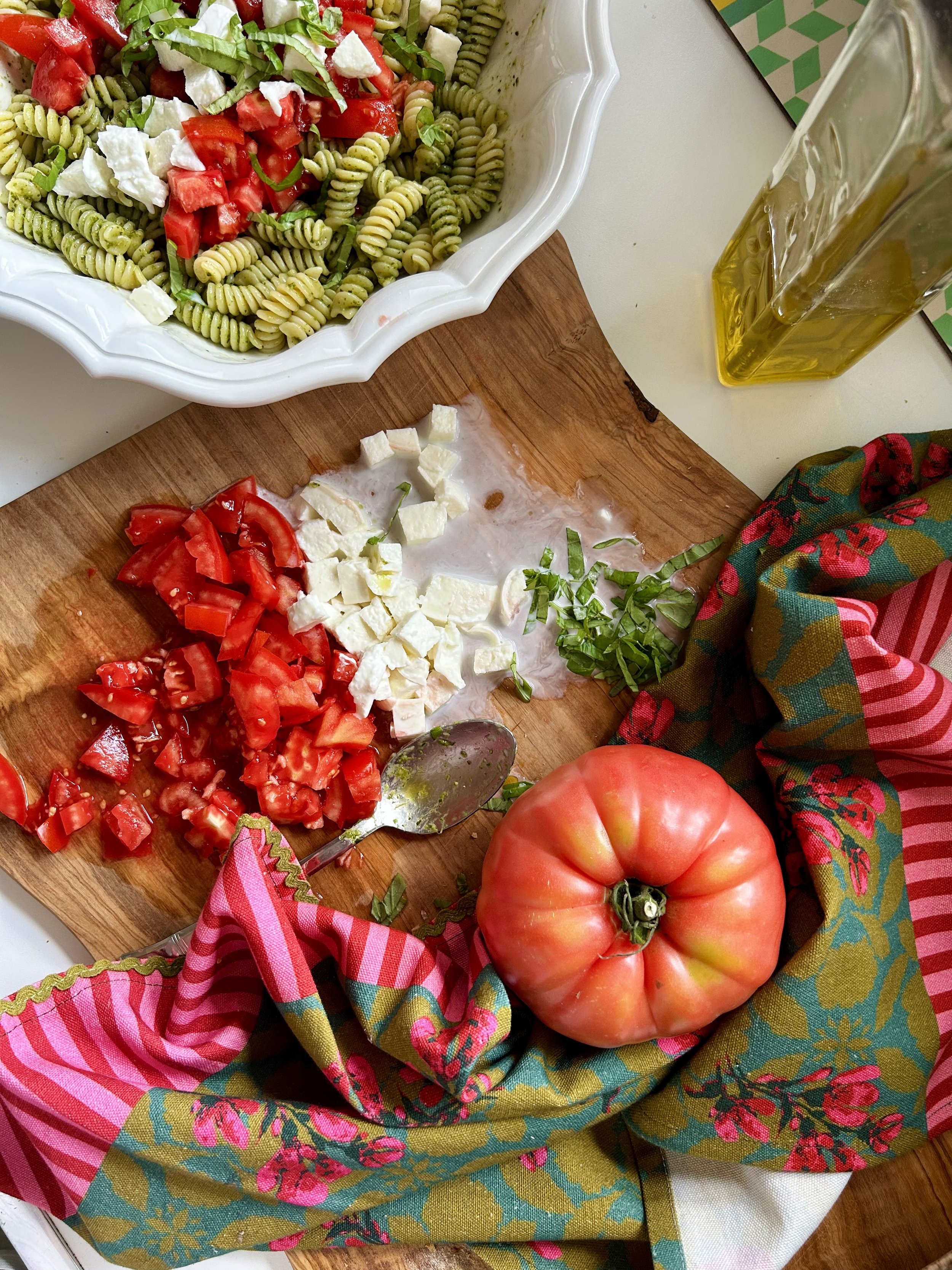 Pink and teal patterned cotton kitchen towel beside a wooden board of chopped tomatoes, feta and basil