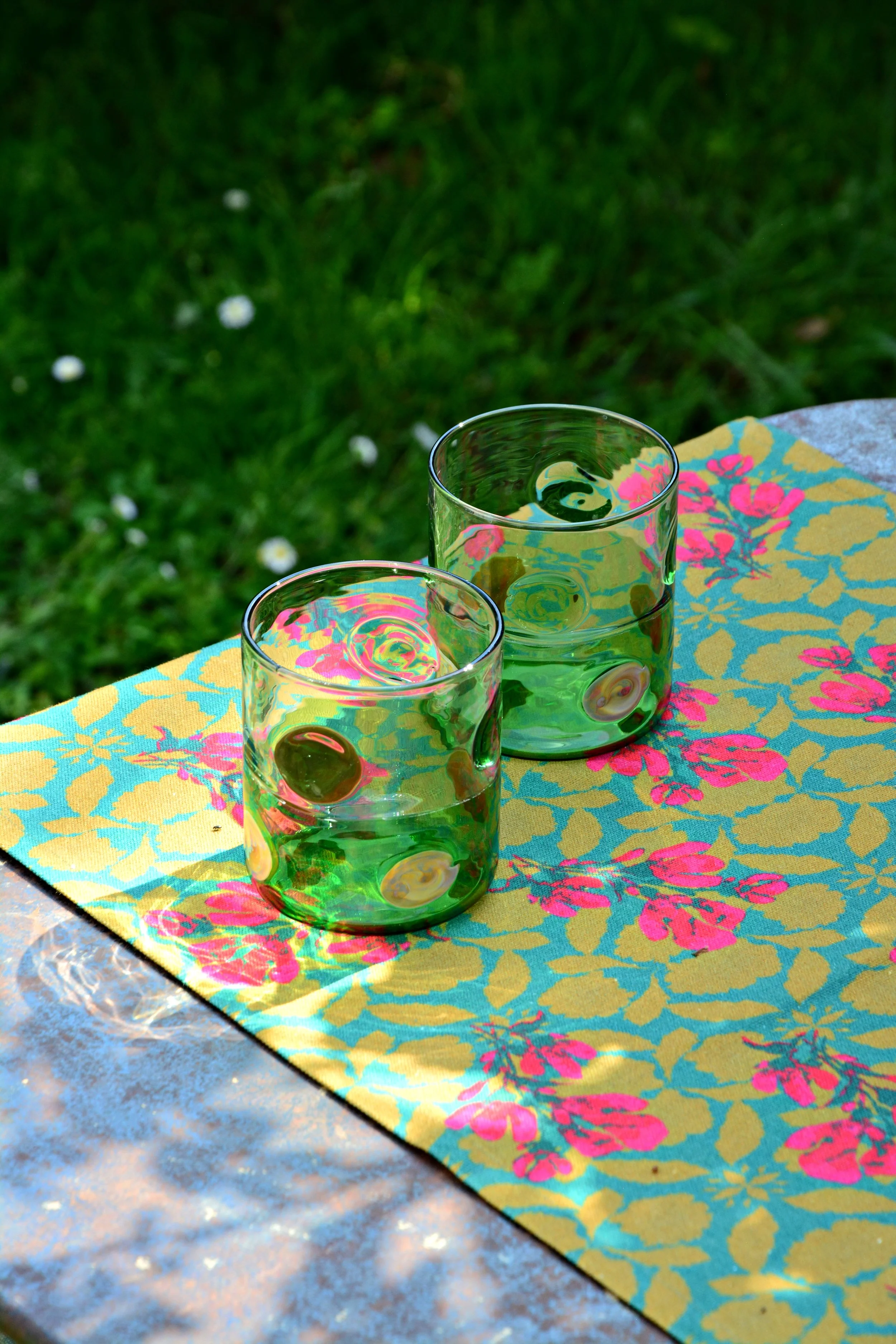 Two transparent glasses filled with a yellowish-green beverage and green olives in a floral-patterned cloth on a table outdoors, with green grass in the background.