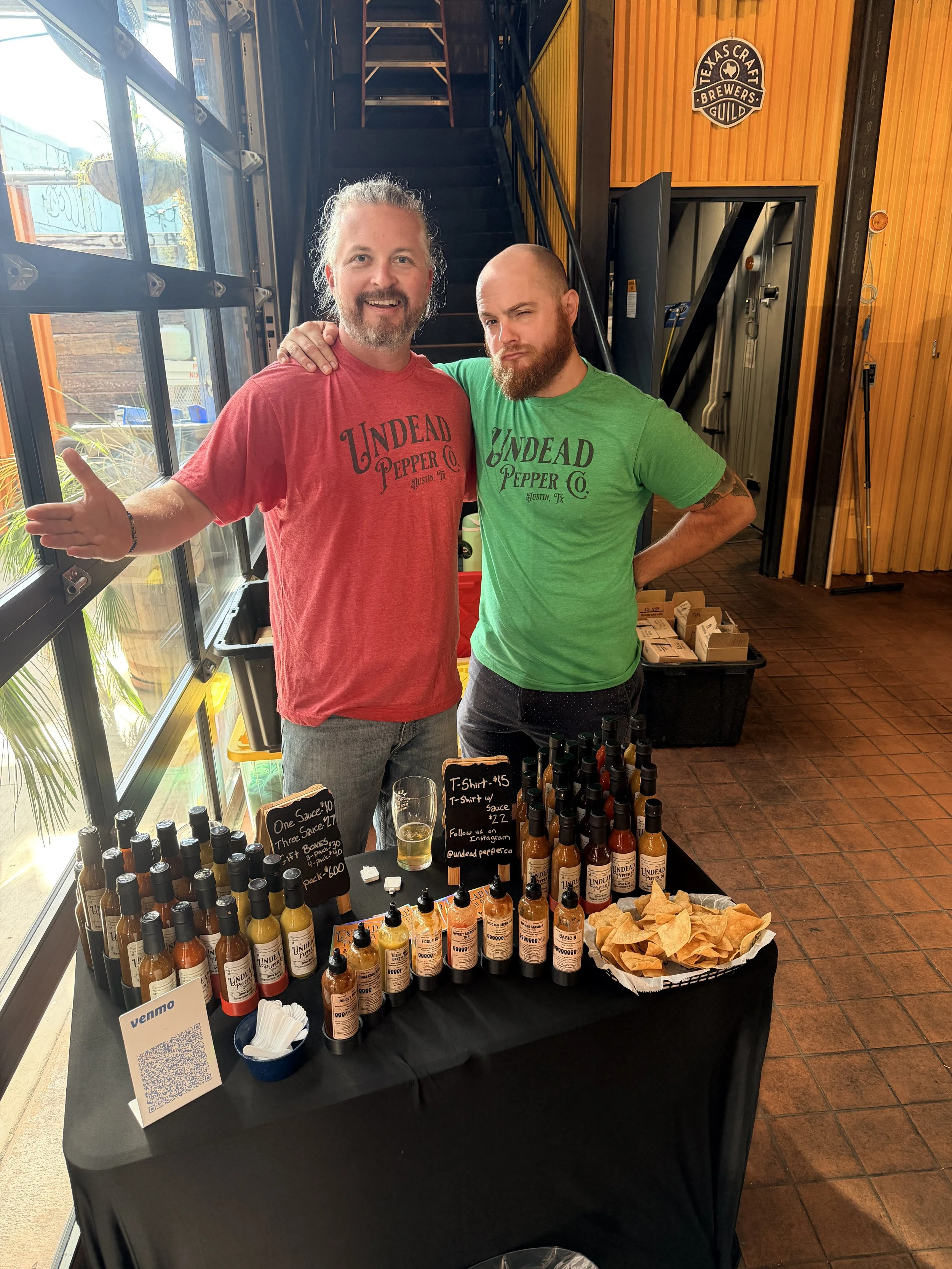 Two men standing behind a table filled with bottles of hot sauce at a vendor booth. One man has long hair and a beard, wearing a red T-shirt, and the other has a tattoo on his arm, a beard, and is wearing a green T-shirt. The booth is set inside a building with wooden walls and a logo that says 'Texas Craft Brewers Guild' visible on the wall.