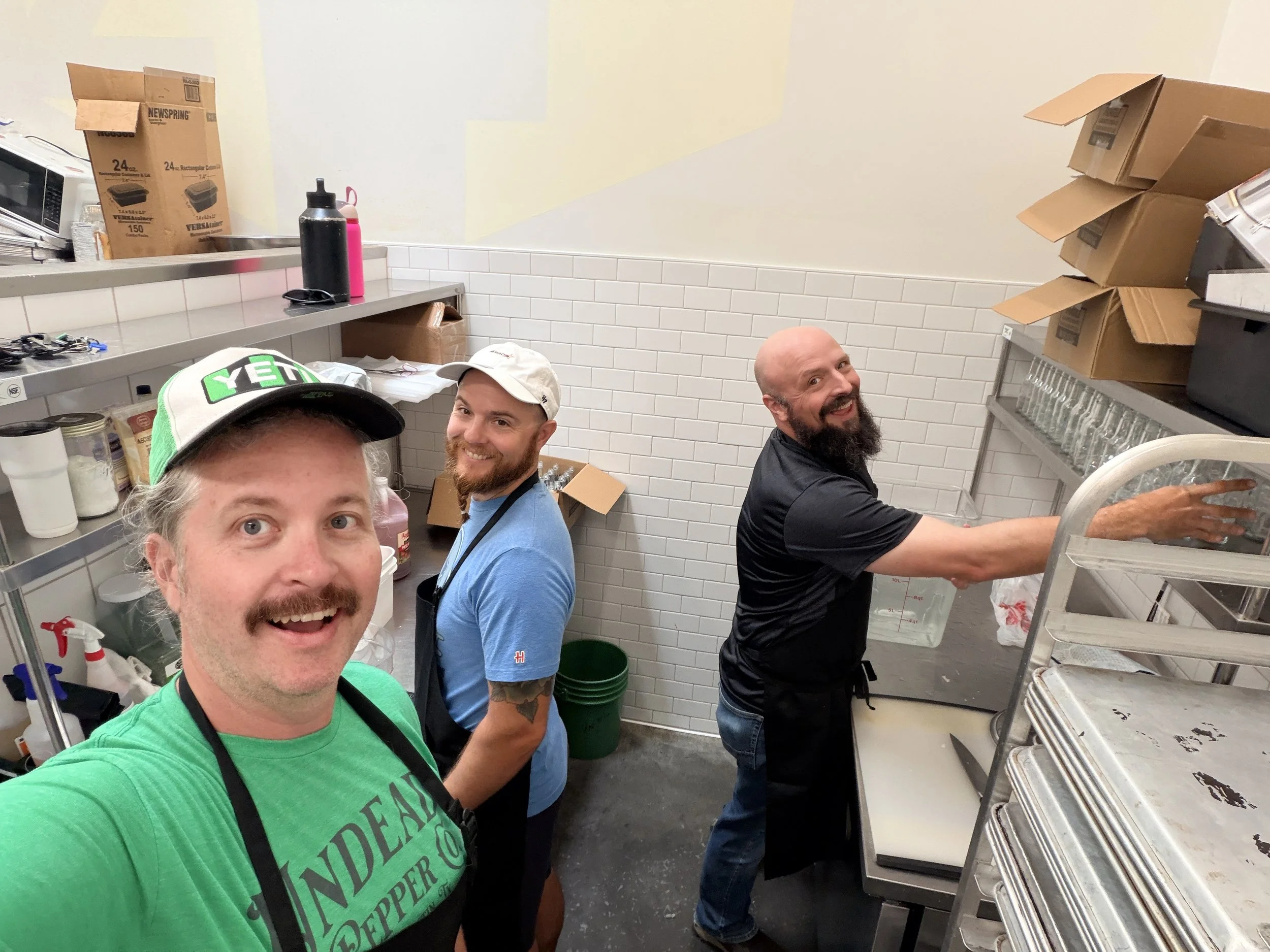 Four men are smiling in a commercial kitchen with boxes and trays. One takes a selfie, and the others appear to be working or preparing food.