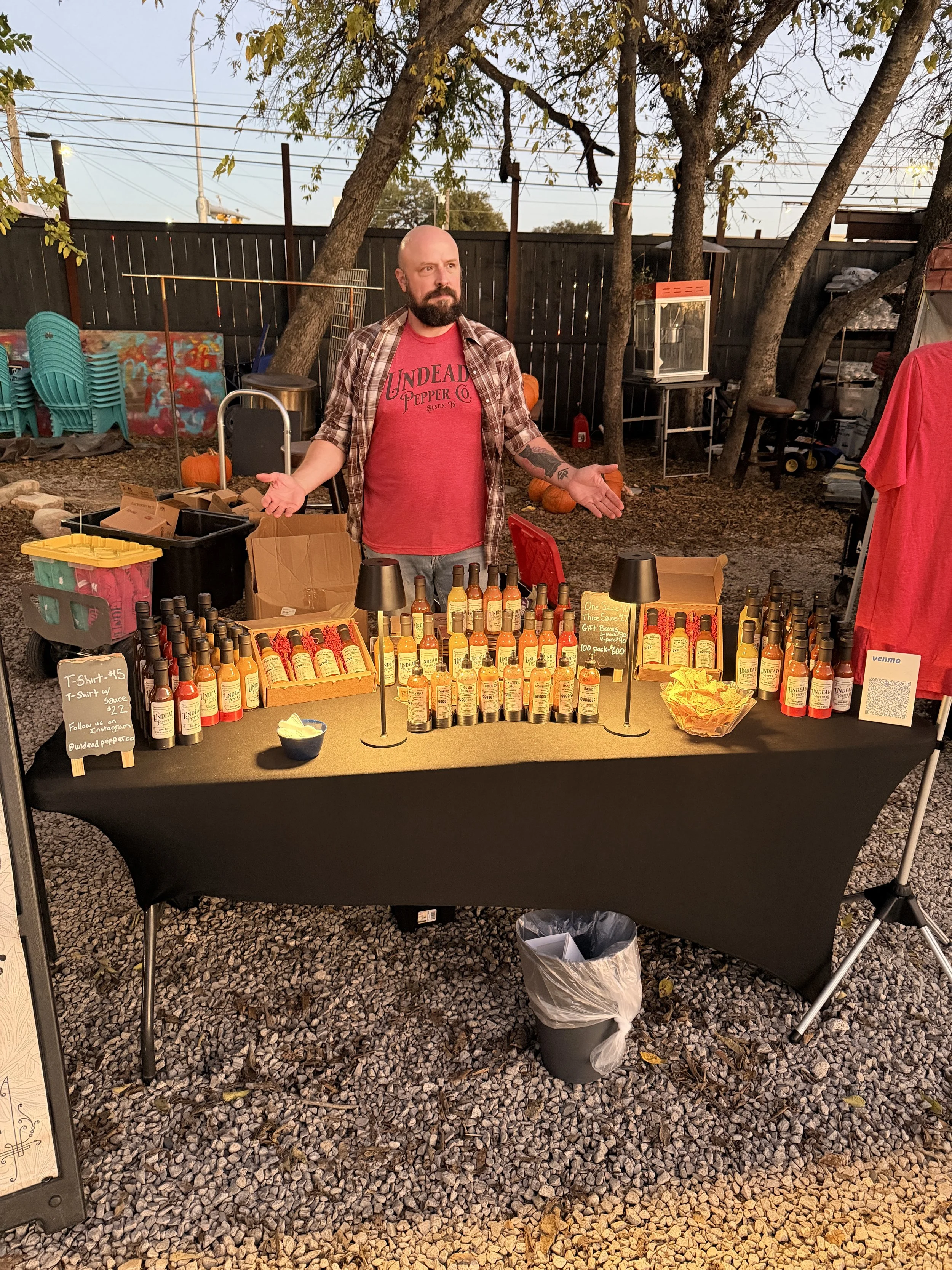 A man with a beard and tattoos standing behind a table of hot sauce bottles at an outdoor market stall during dusk. The table displays various bottles, and there are pumpkins and fall decorations around. The man is wearing a red T-shirt and a plaid shirt.