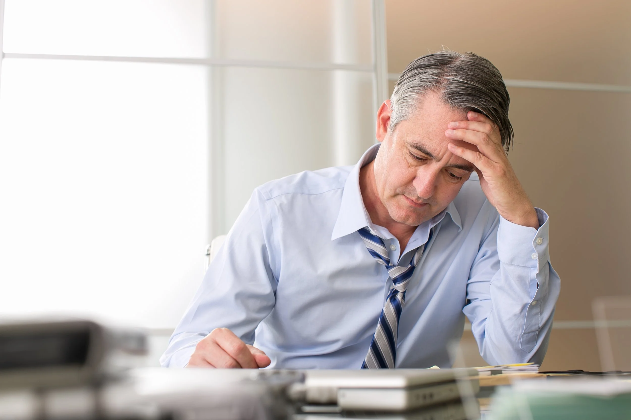 A middle-aged man wearing a light blue dress shirt and a striped tie, sitting at a desk, holding his forehead with one hand, appearing stressed or worried.