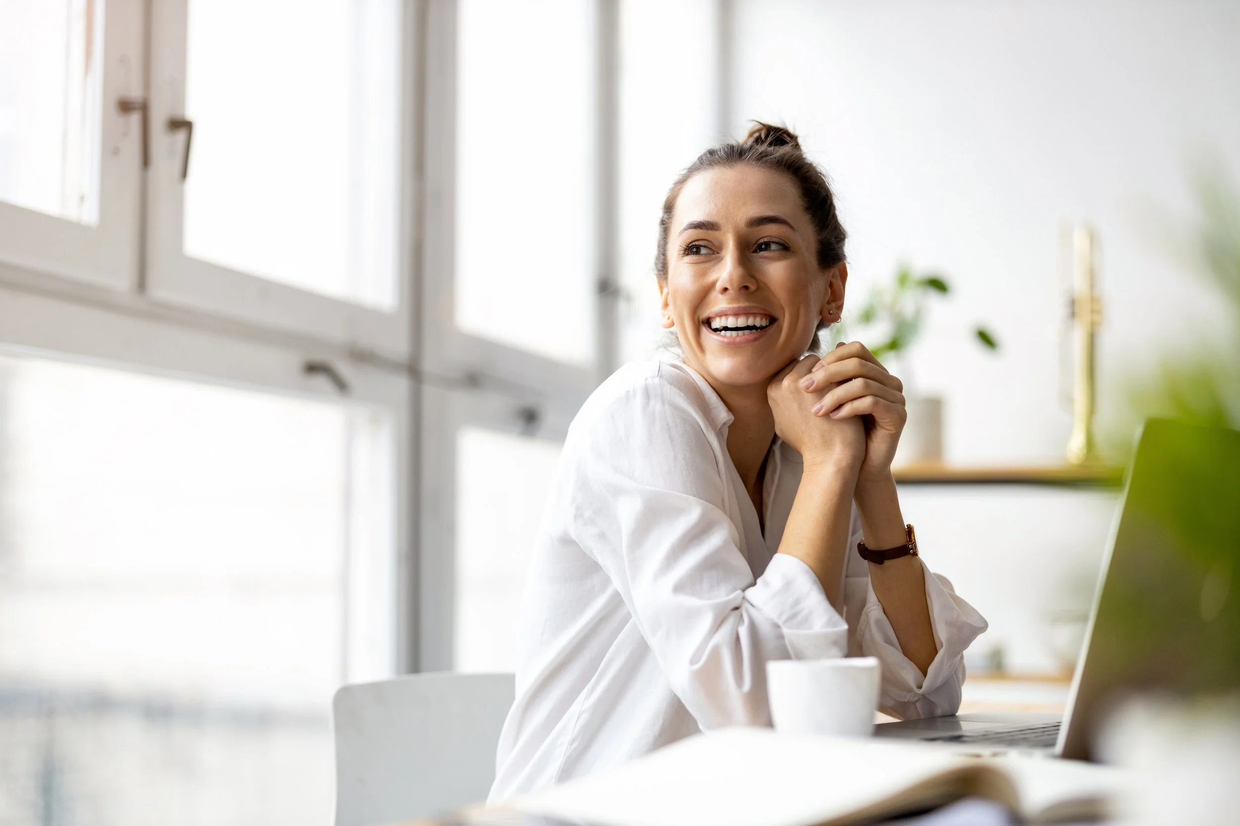 A young woman with brown hair tied in a bun, smiling and looking to the side, sitting at a desk with a laptop, in a bright room with large windows and some plants in the background.