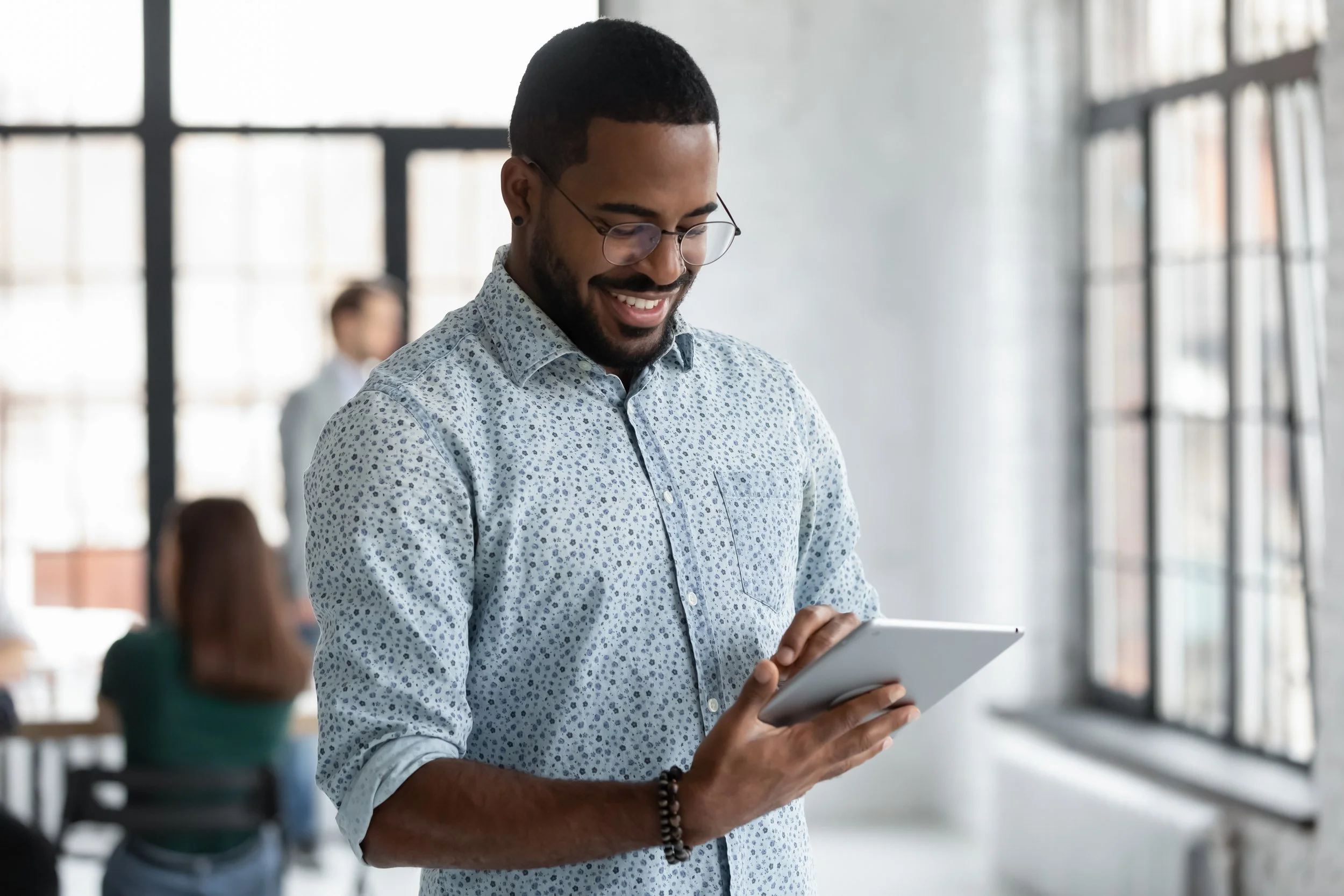 A man with glasses and a beard wearing a white shirt with a blue pattern, looking at a tablet and smiling, in a modern office space with large windows and other people in the background.