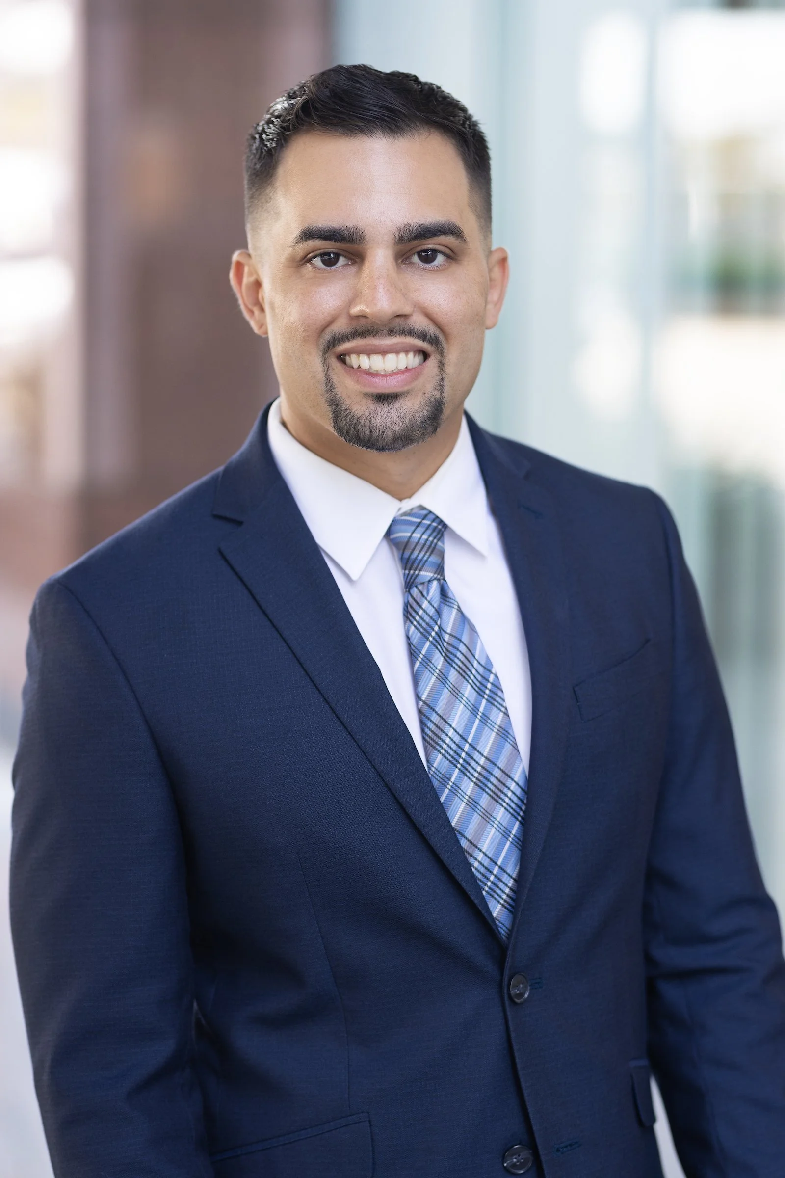 A man with short dark hair, a goatee, and a mustache, dressed in a navy blue suit, white shirt, and a blue and white plaid tie, standing in an office with a blurred background and bright natural lighting.