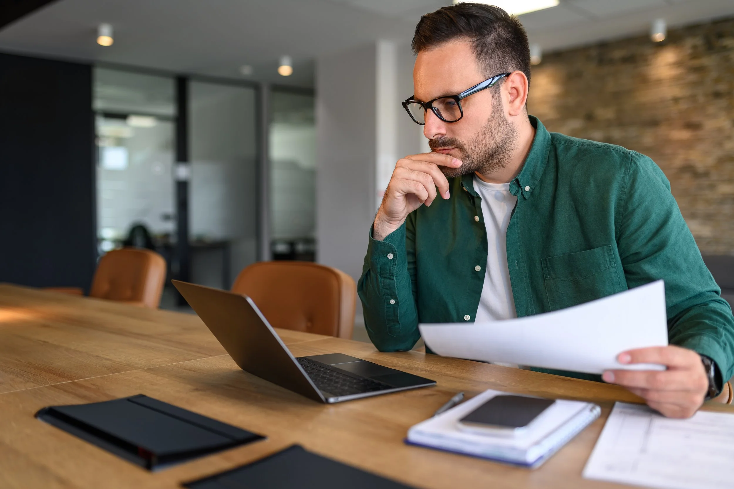 Man with glasses sitting at a wooden table, looking at a smartphone in one hand, with a laptop and papers in front of him in a modern office.