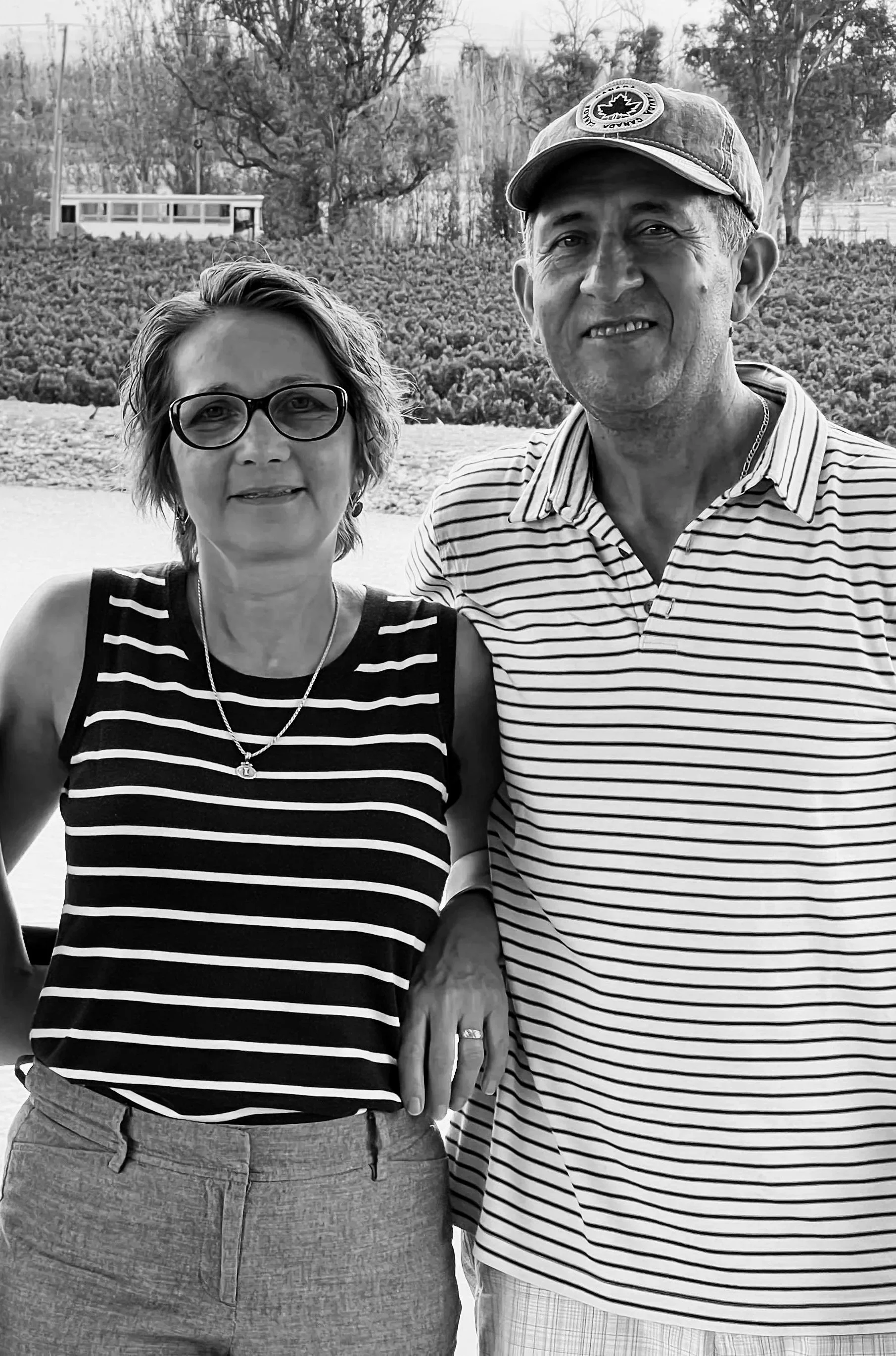 A black and white photo of a woman and a man standing outdoors, holding hands. The woman has short hair, glasses, and is wearing a sleeveless striped top and a necklace. The man has short hair, is smiling, and is wearing a striped polo shirt, a cap with a logo, and a chain necklace. They are in a park or garden with trees and shrubbery in the background.