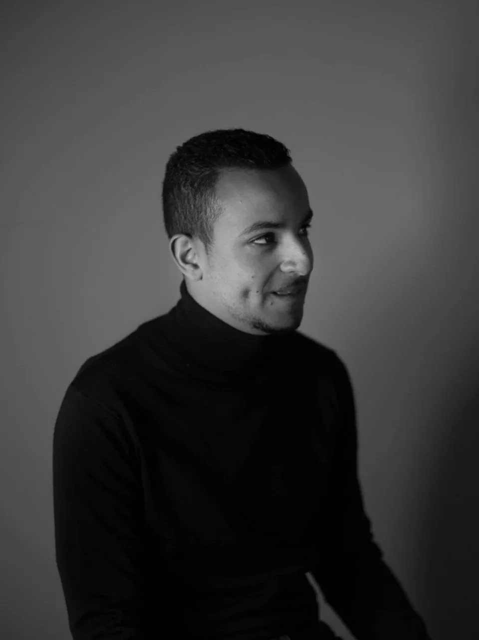 Black and white portrait of a man in a turtleneck, looking to the side with a subtle smile.