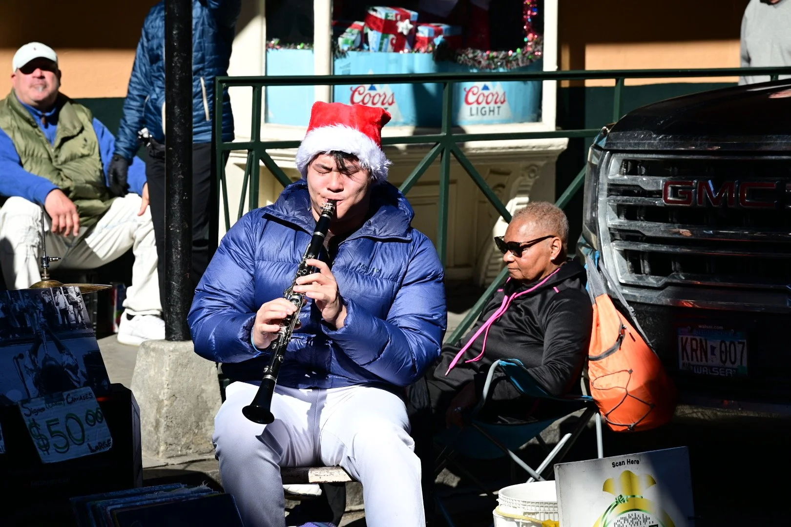 A man wearing a Santa hat and a blue jacket is playing the clarinet outdoors during a holiday event. Sitting nearby are two people, one wearing sunglasses and a black jacket, and the other in the background with a green vest and white pants. There's a decorated storefront with Christmas presents and a Coors Light sign, and a GMC vehicle is parked beside them.