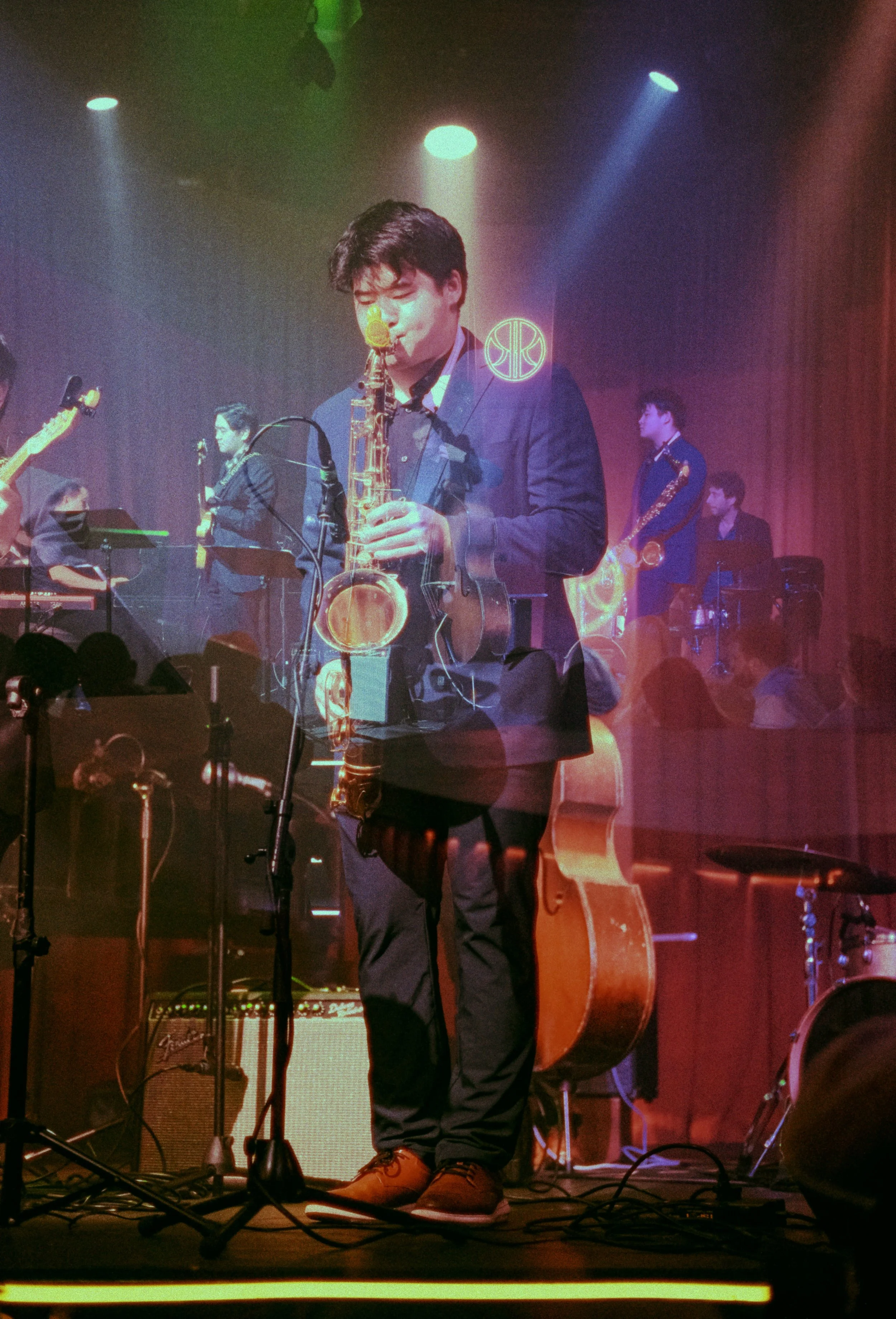 Musician playing the saxophone on stage with a guitar and drums in background, colorful stage lights.