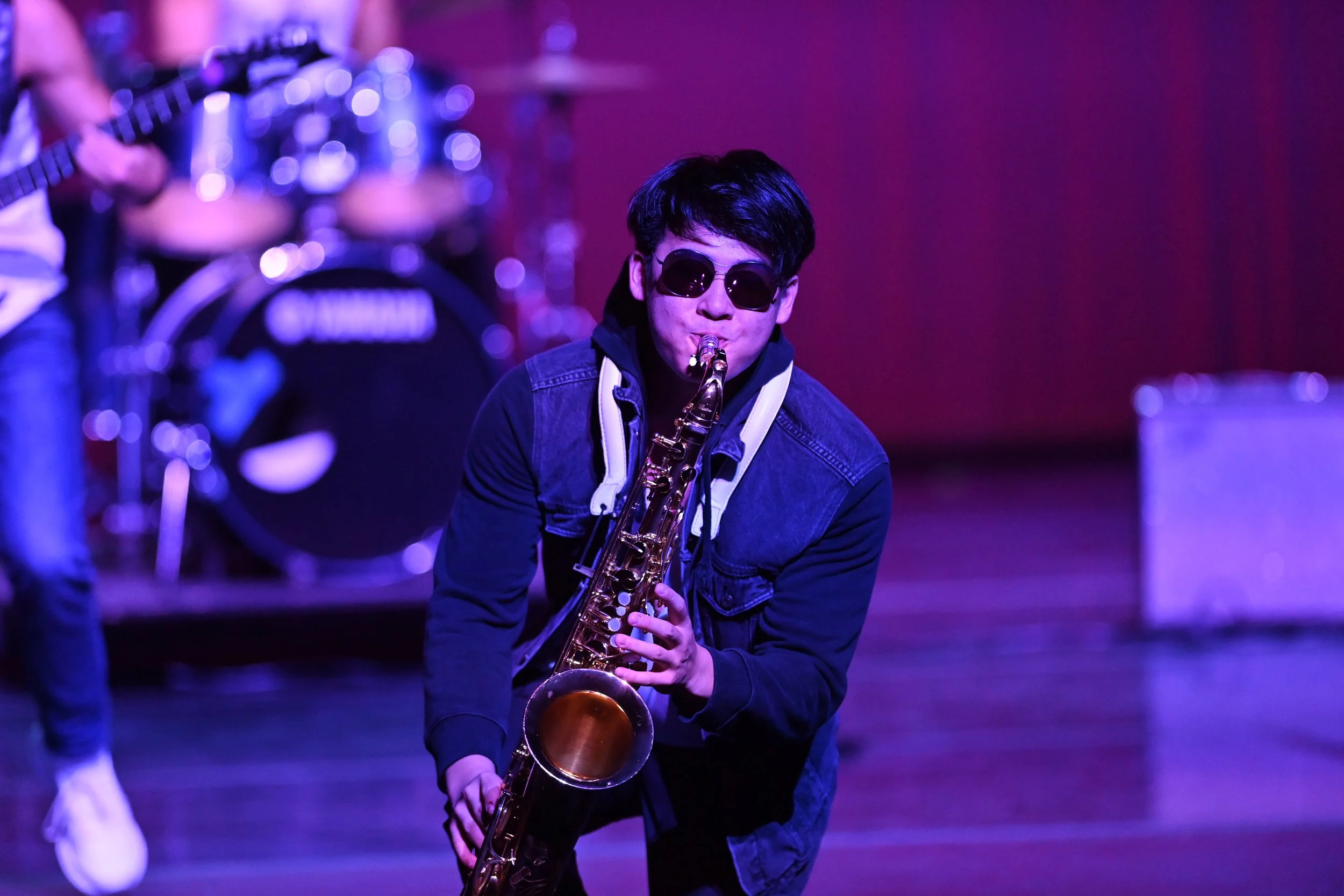 A young man with black hair wearing sunglasses, a denim jacket, and headphones is playing a saxophone on stage with colorful stage lighting.