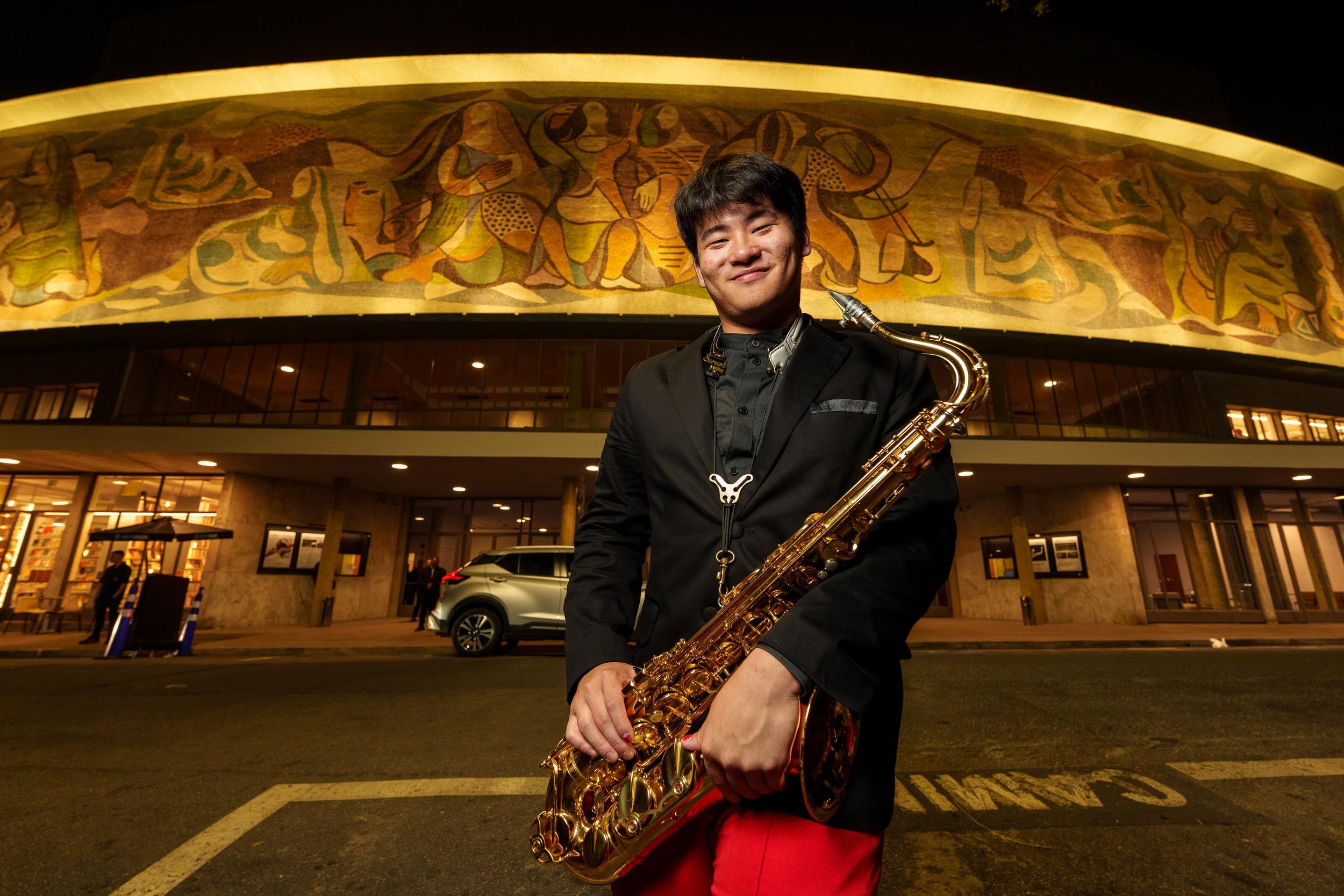 A young man dressed in black with red pants holding a gold saxophone in front of a large mural at night.