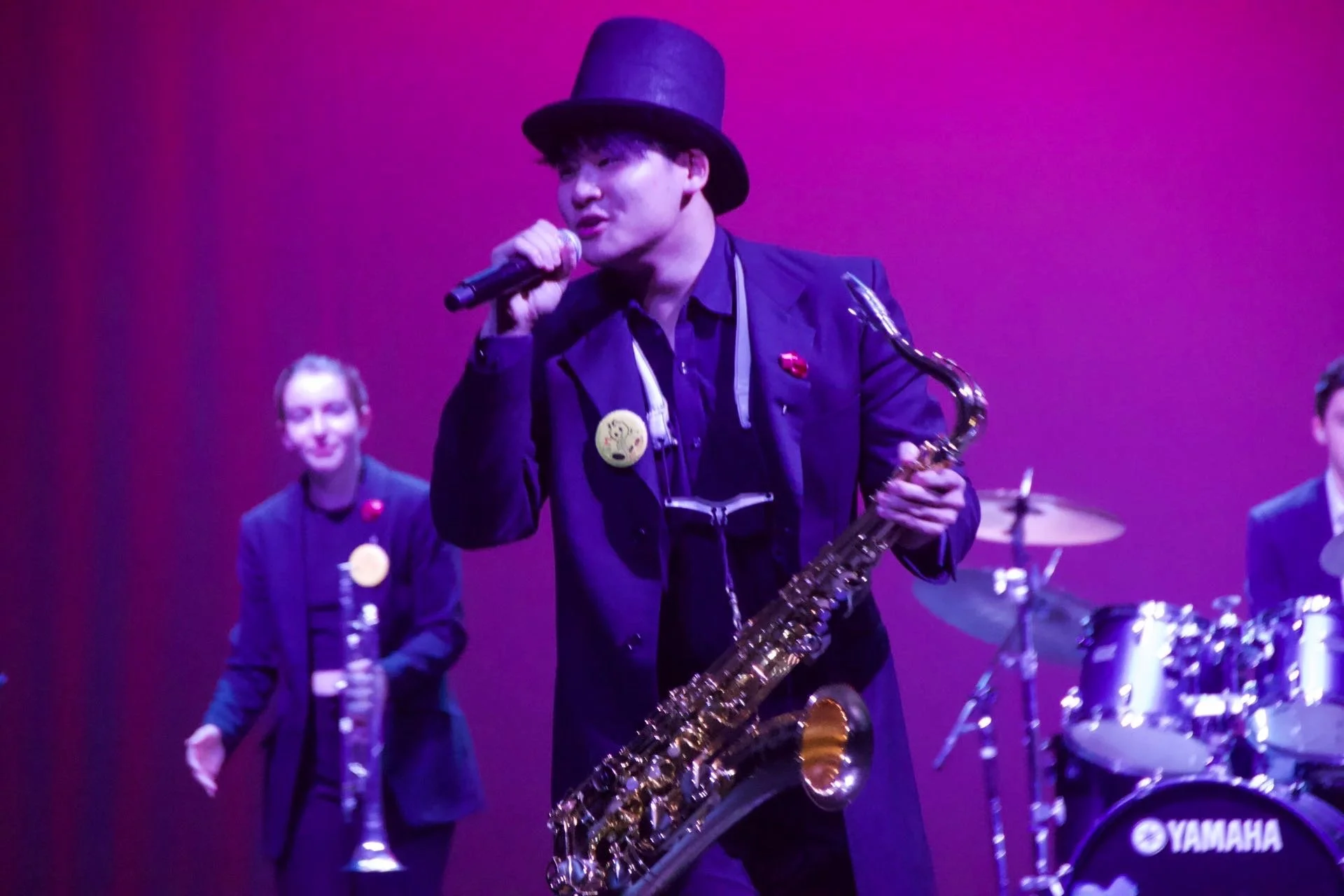 A man wearing a black hat, black jacket, and a red pin, holding a saxophone and singing into a microphone on a stage with purple lighting, accompanied by two women and a drum set.