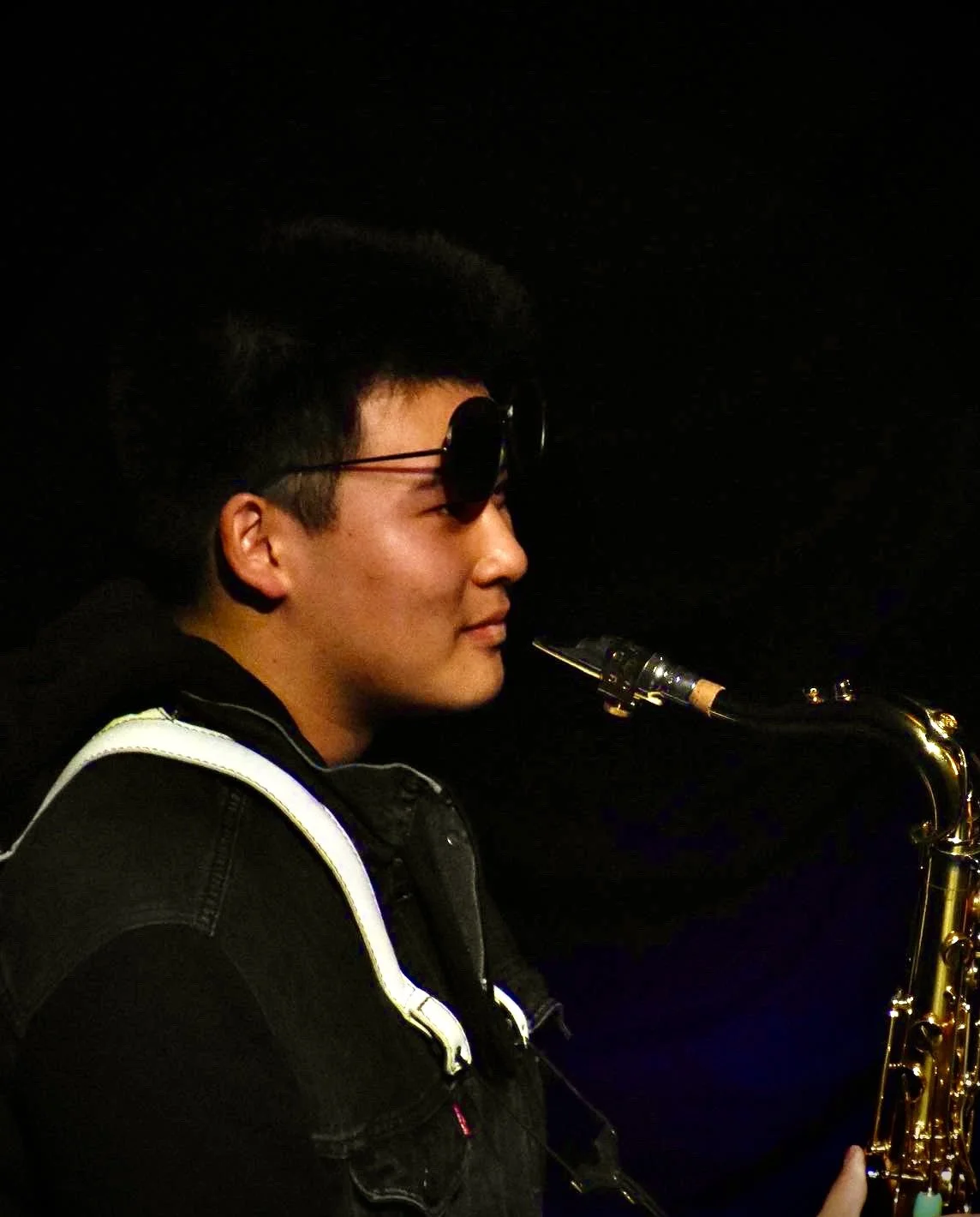 Young man playing a saxophone at night, wearing sunglasses on his head and a black vest.