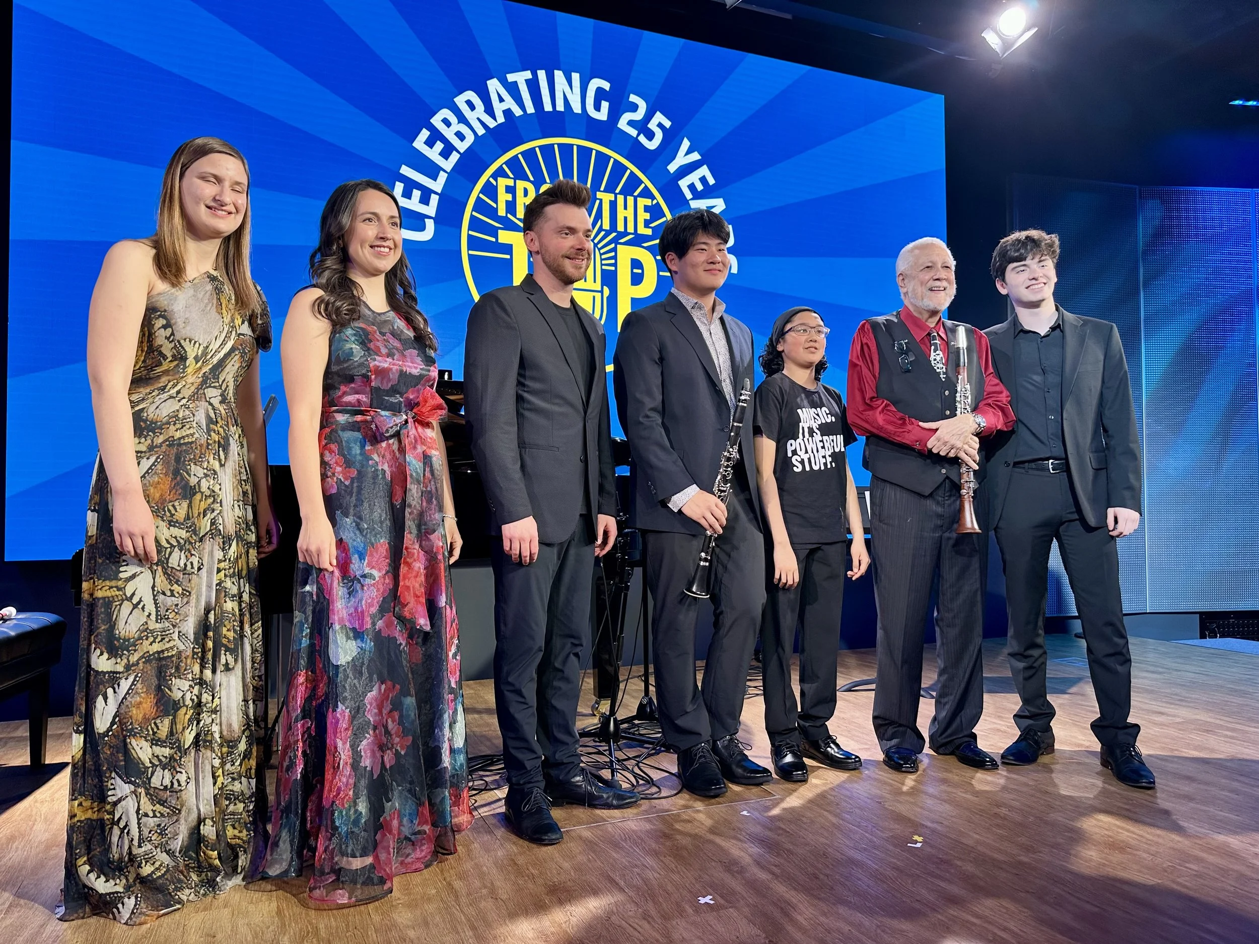 Group of six people standing on stage in front of a large blue screen that reads 'Celebrating 25 Years of the' with part of a yellow graphic logo. The group includes three women, two men, and a child, all dressed in formal or semi-formal attire.