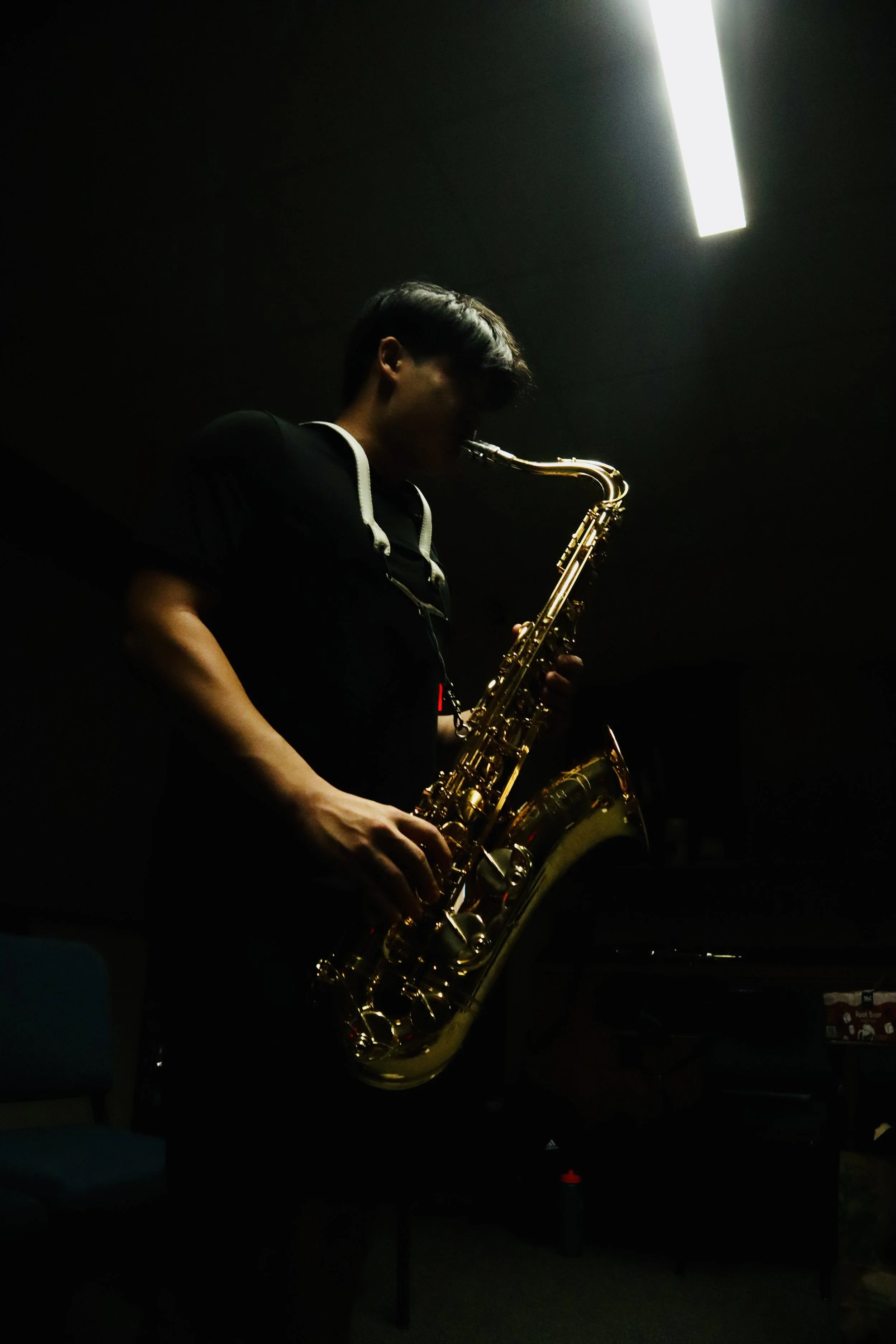 A person playing a golden saxophone in a dark room, illuminated by a single bright light overhead.