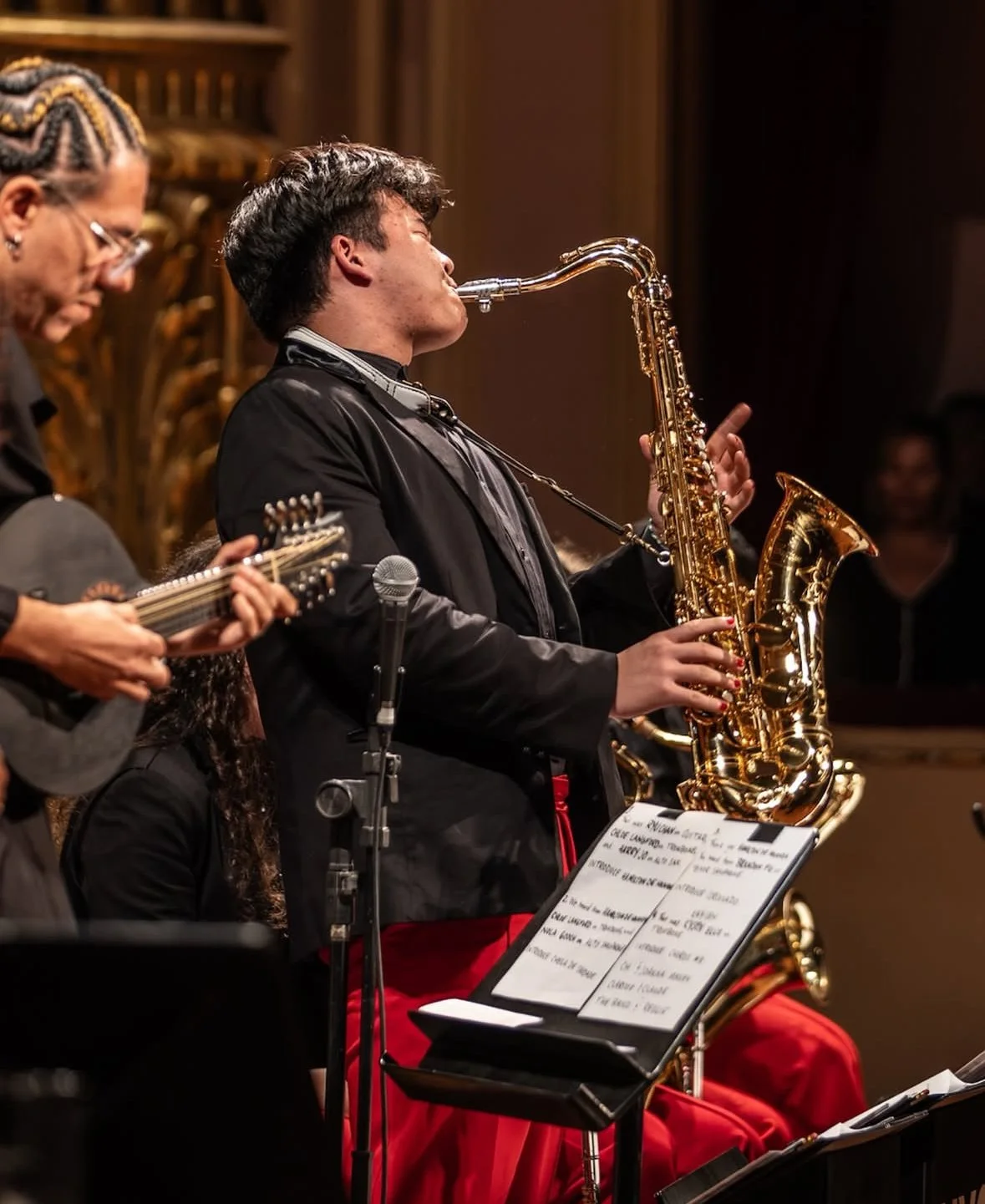 A musician playing a saxophone on stage during a performance, with sheet music on a stand in front.