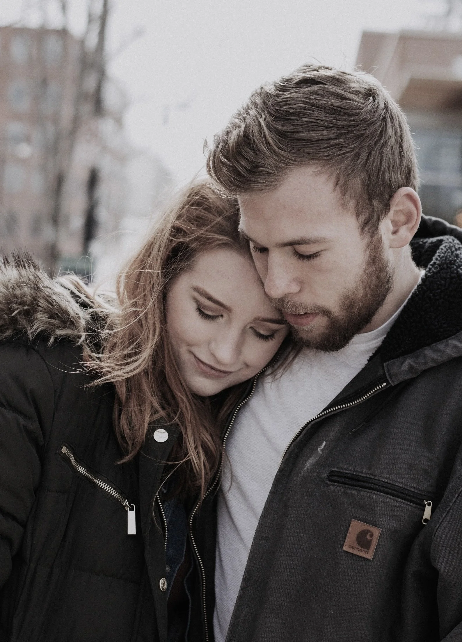 A close-up of a young man and woman with their heads resting against each other, eyes closed, and smiling softly in an outdoor setting.