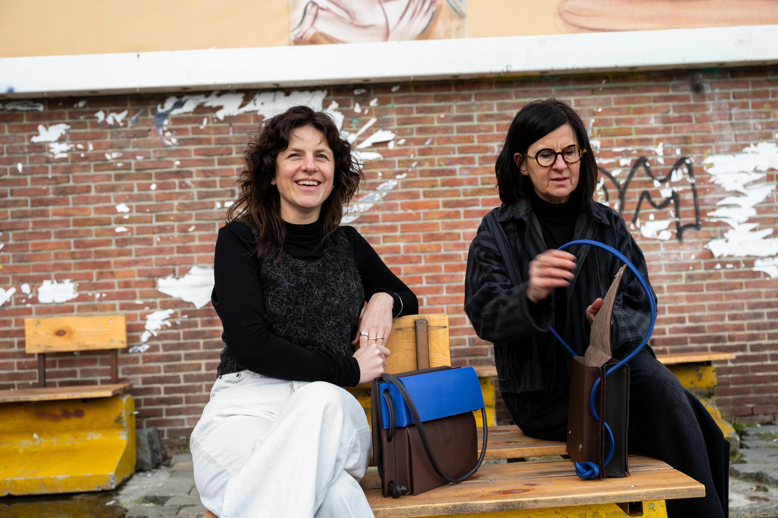 Sien en Annemie zitten op een bank aan Dok-Noord in Gent. Beide hebben ze een handtas van Jacqo bij. Ze hebben verschillende vorm, kleur.