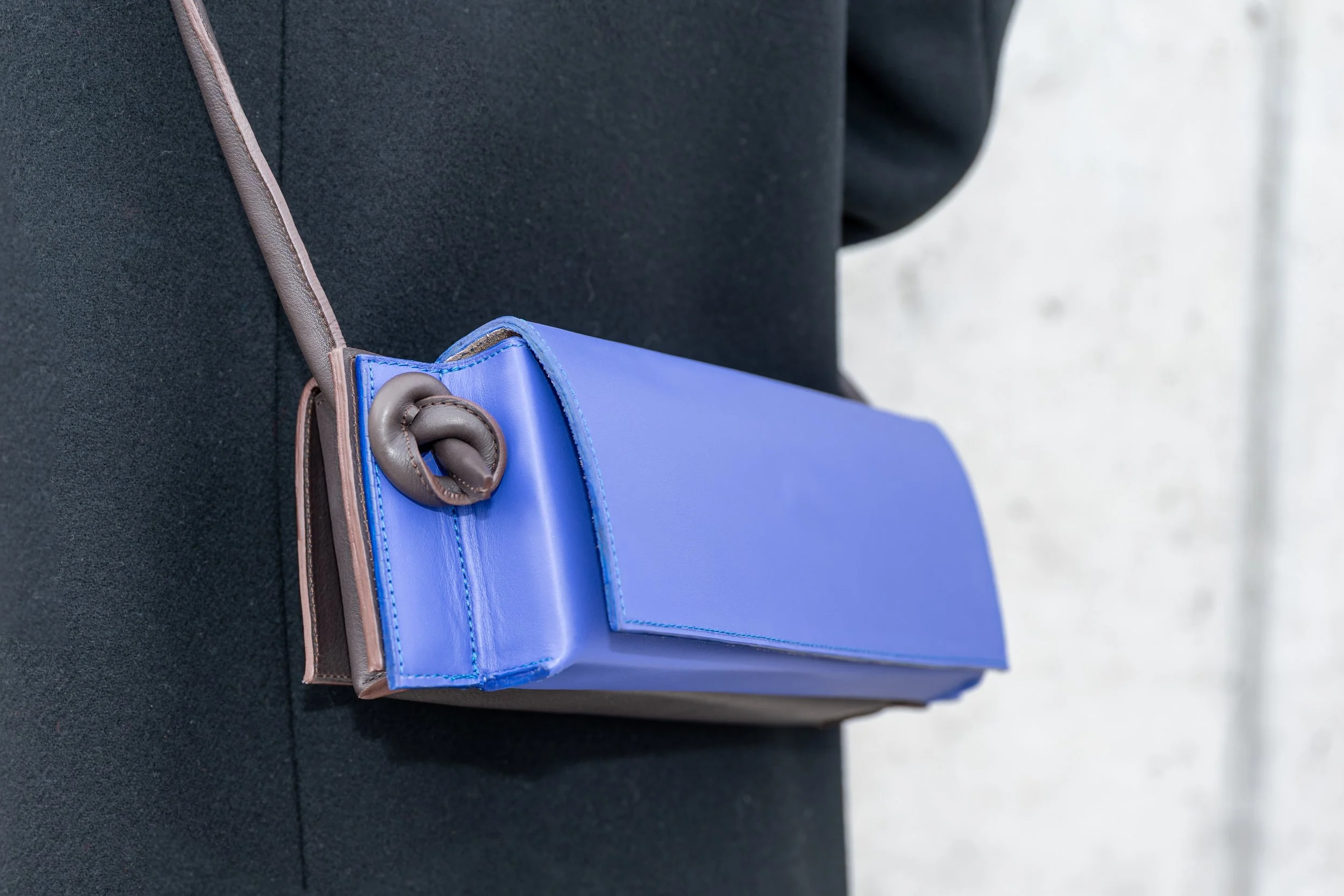 Een blauwe handtas met bruin leren details hangt aan de schouder van iemand die een zwarte jasje draagt.