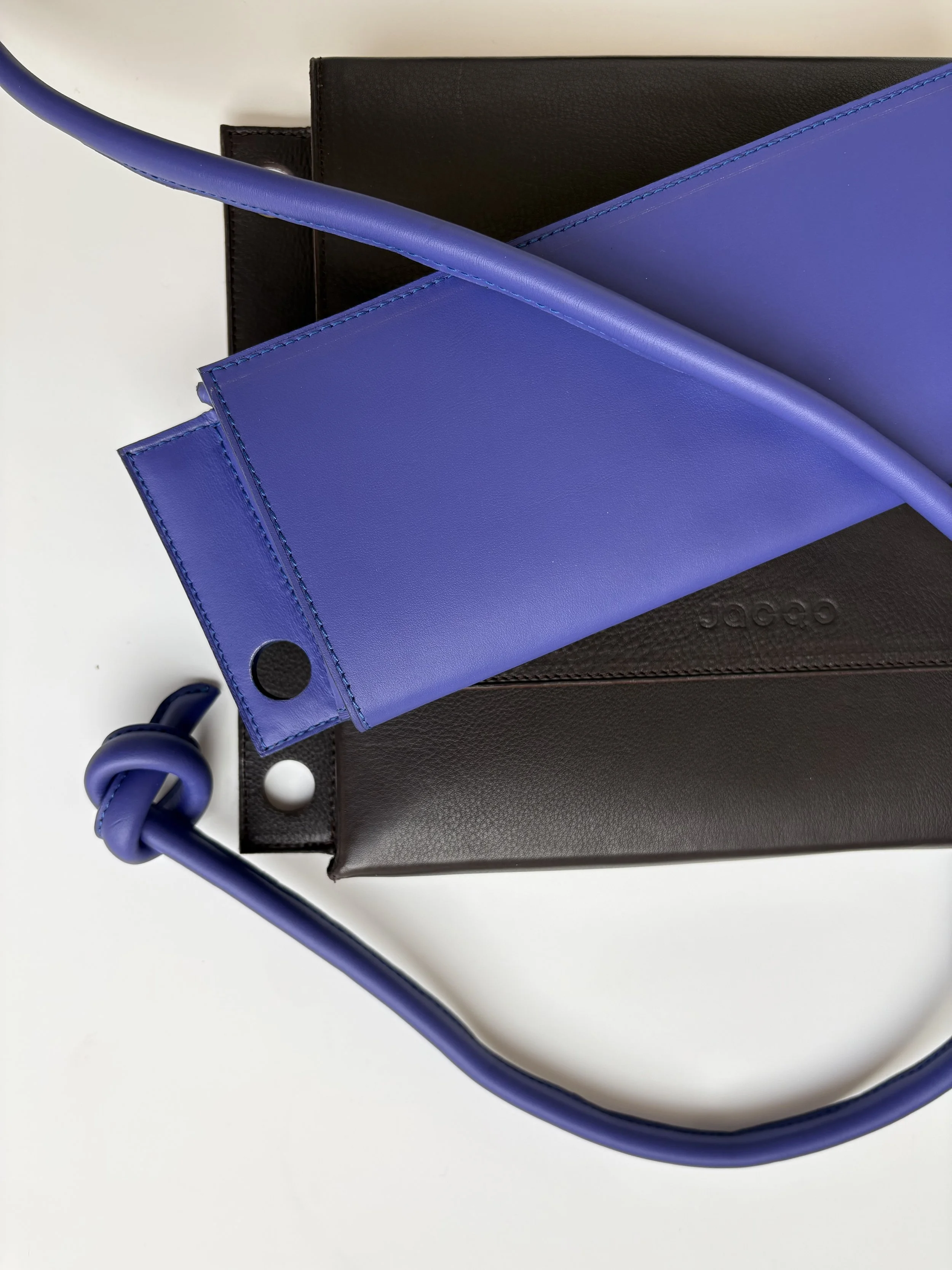 Drie kleurrijke handtassen in blauw, zwart en bruin liggen op een witte achtergrond. Boven de handtassen is een donkerblauw lint, de draagriem, te zien. Er zit een knoop in de draagriem.