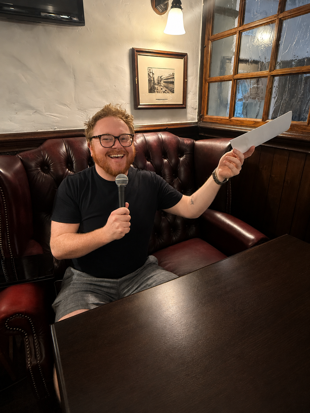 A man with glasses and a beard sitting on a leather booth in a restaurant, holding a microphone and a piece of paper, smiling at the camera.