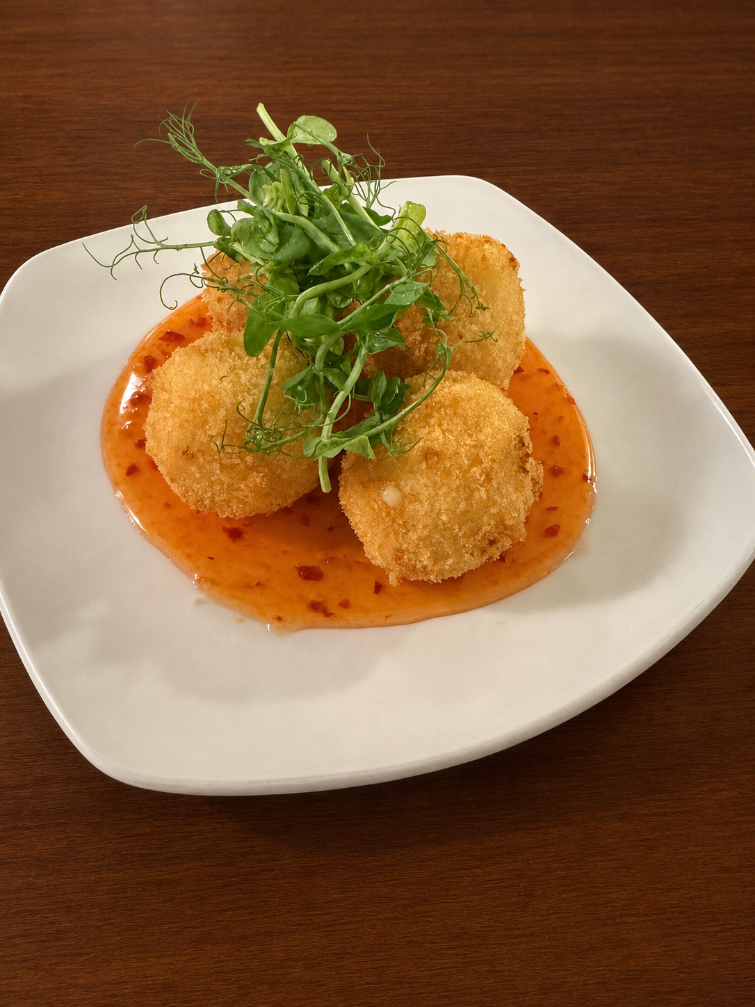 Crispy fried croquettes topped with microgreens, served on a bed of orange sauce with chili flakes in a white dish on a wooden table.
