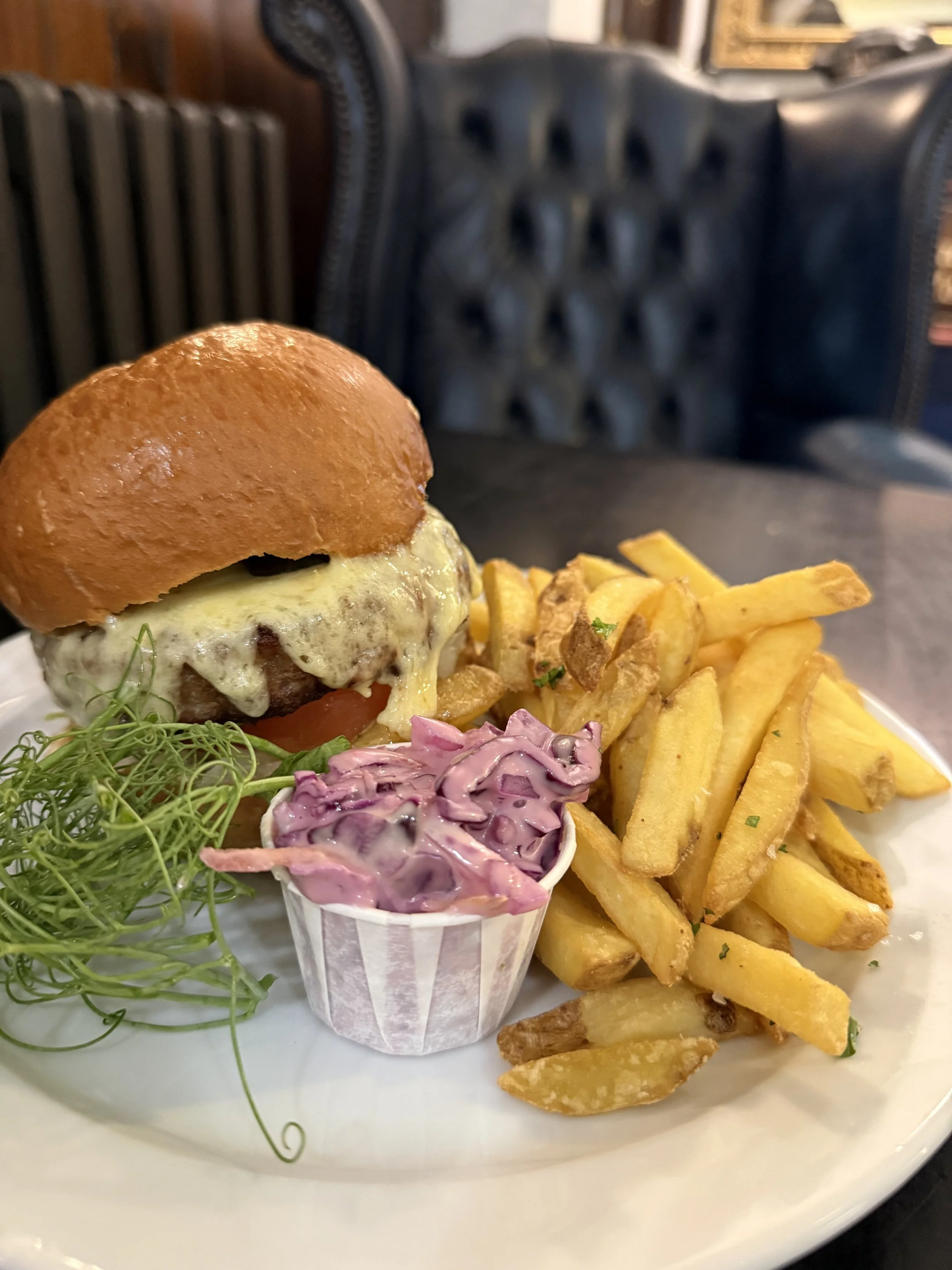 A cheeseburger with lettuce, tomato, and melted cheese, served with a side of French fries and coleslaw, garnished with a sprig of greens, on a white plate.