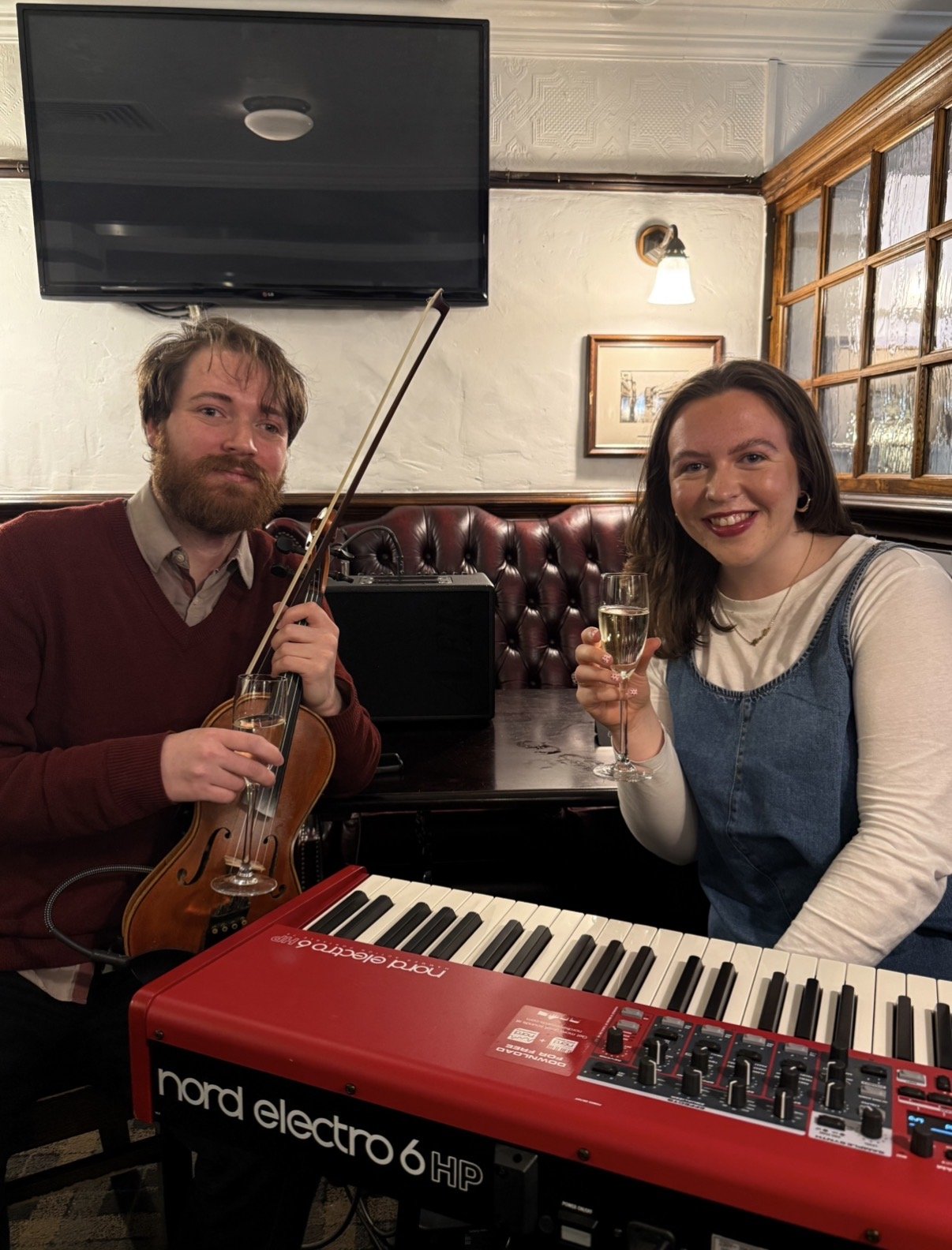 A man with a beard playing a violin and a woman smiling with a glass of champagne sitting at a piano in a cozy room with a TV and wall decorations.