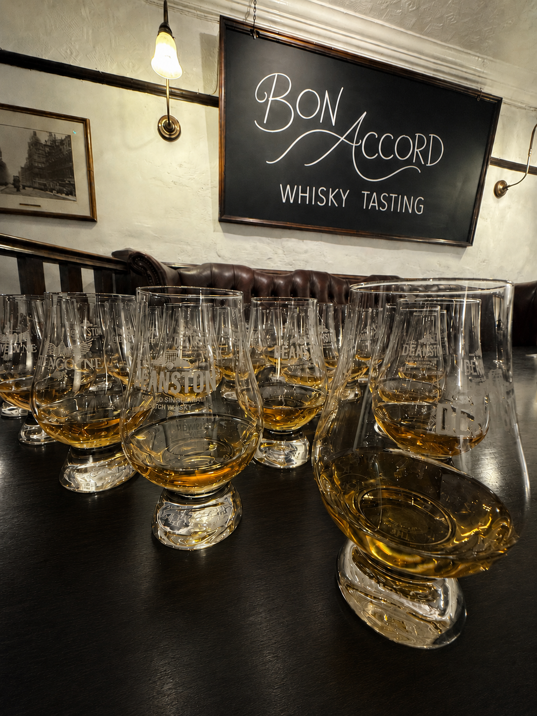 Multiple whisky glasses on a table at a whisky tasting event, with a sign in the background that reads 'Bon Accord Whisky Tasting'.