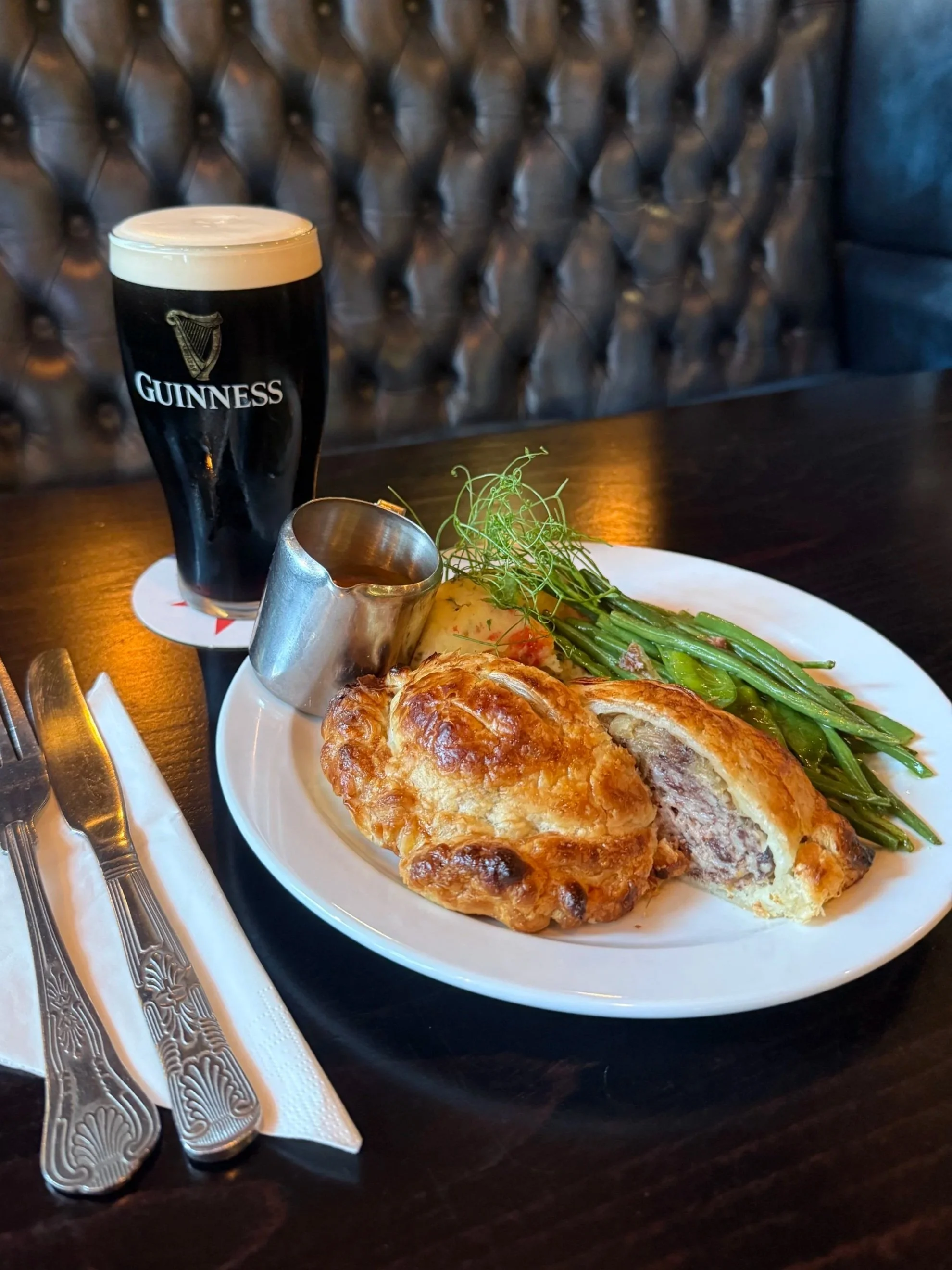 A pint of Guinness beer with a creamy head on a dark table, a white plate with a large Yorkshire pudding stuffed with meat and gravy, green beans, and a garnish of greens. Silverware wrapped in a decorative napkin is placed on the side.