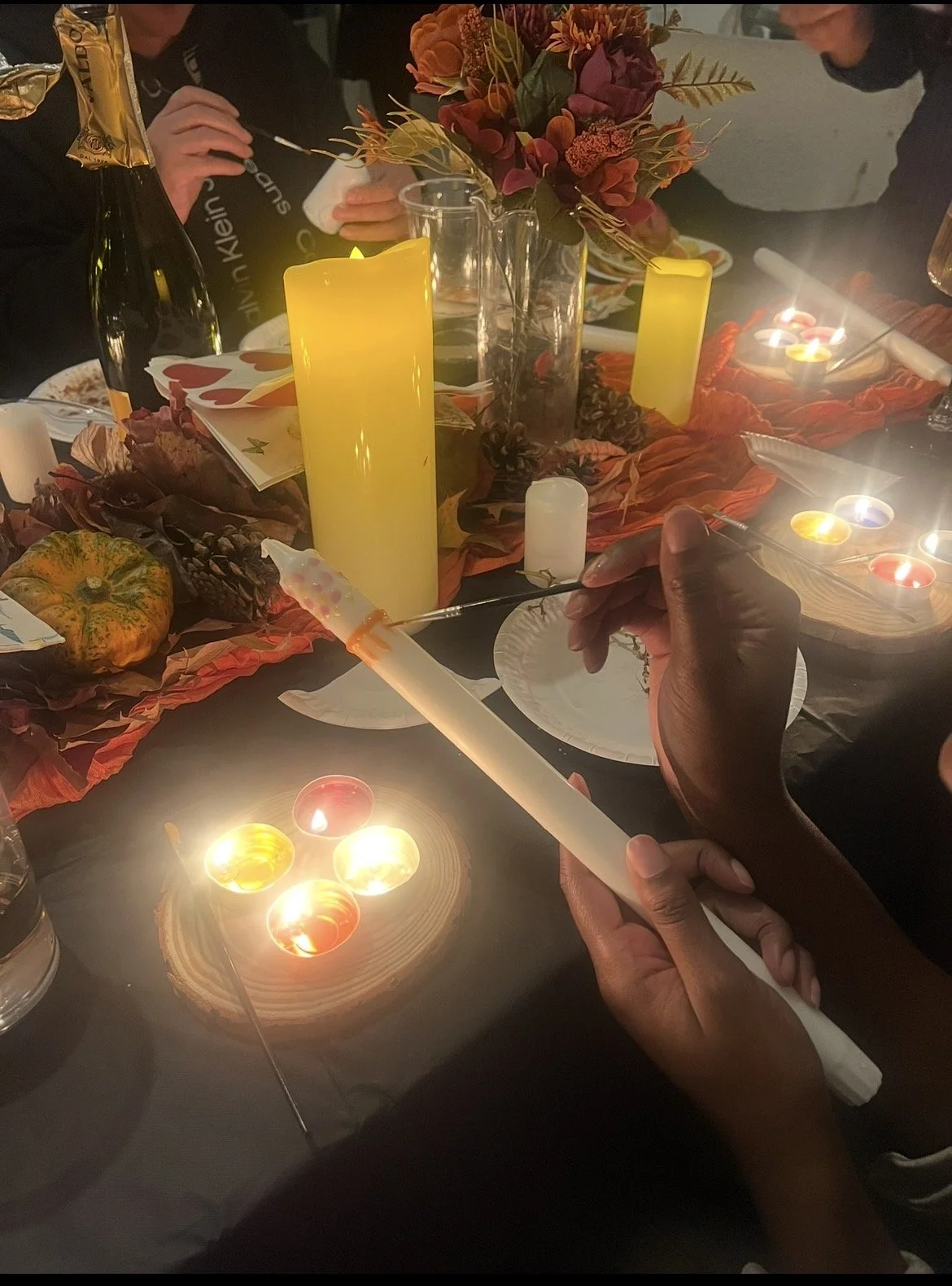 Guests painting taper candles by candlelight at a cosy autumn-themed workshop, surrounded by pumpkins, pine cones and warm floral decor.