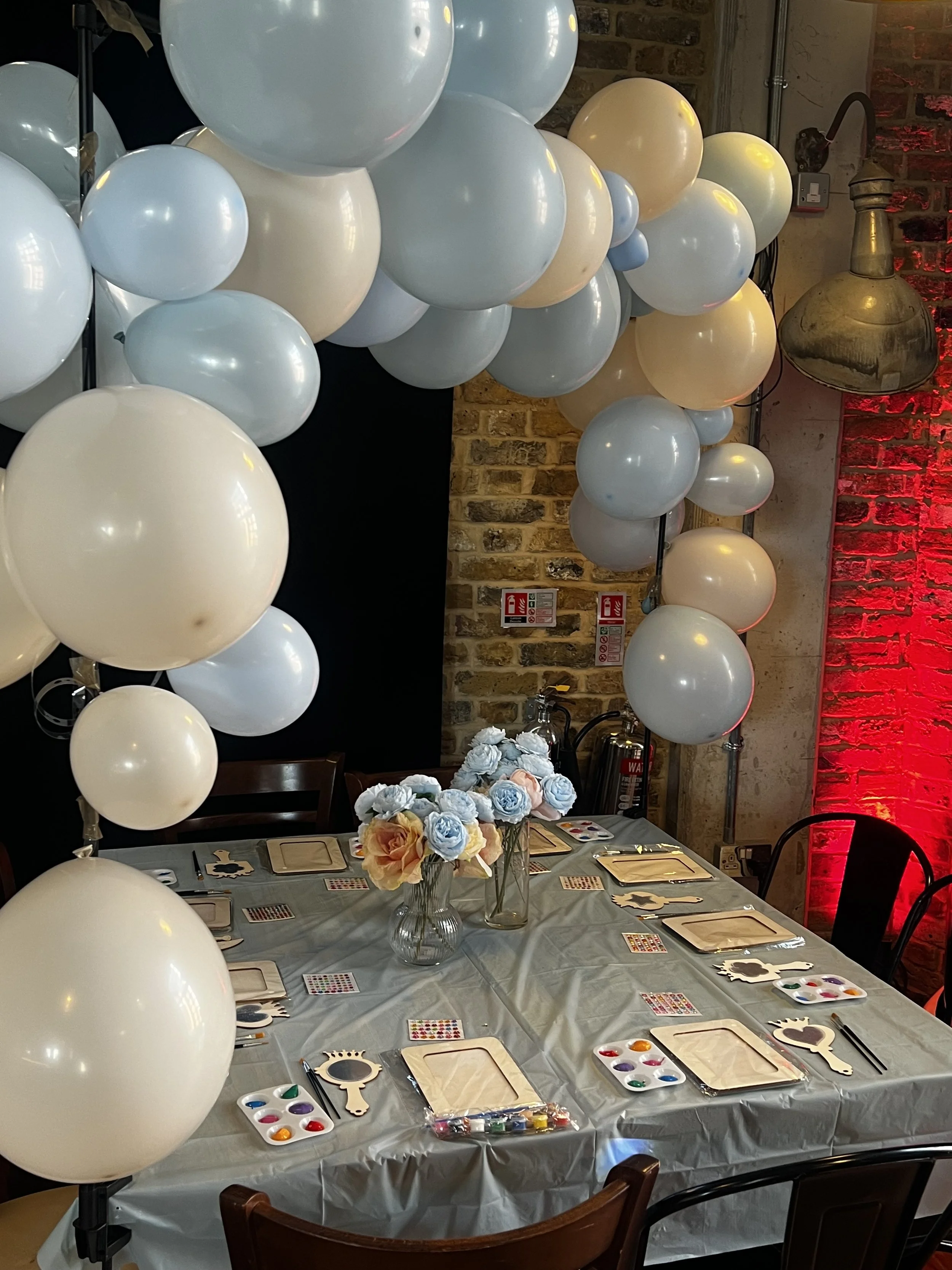 Styled craft party table with wooden frames, paint palettes and balloon arch in blue and white, set in a London brick venue.