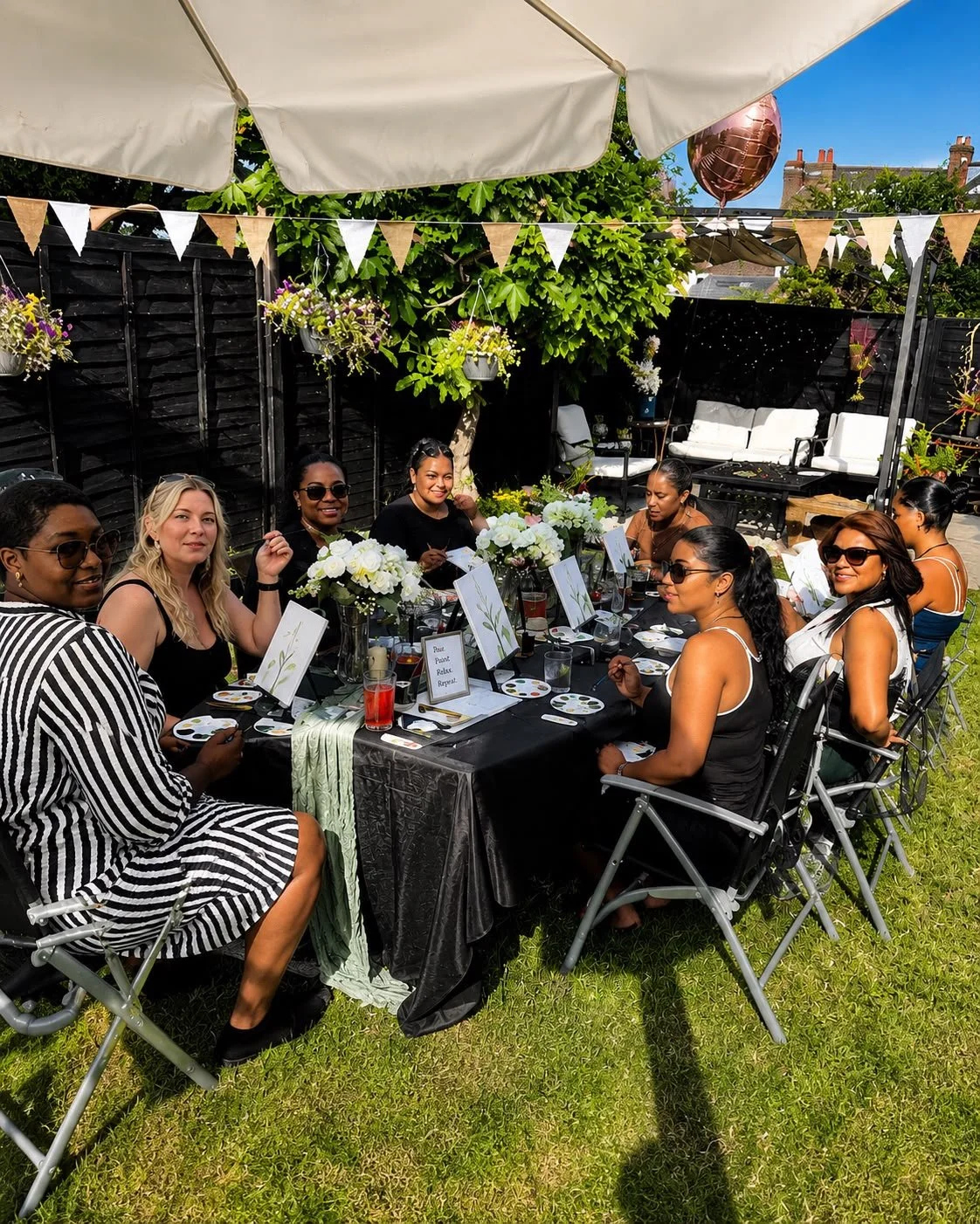 A diverse group of women enjoying an outdoor paint and sip art session at a styled garden table beneath a canopy on a sunny day.