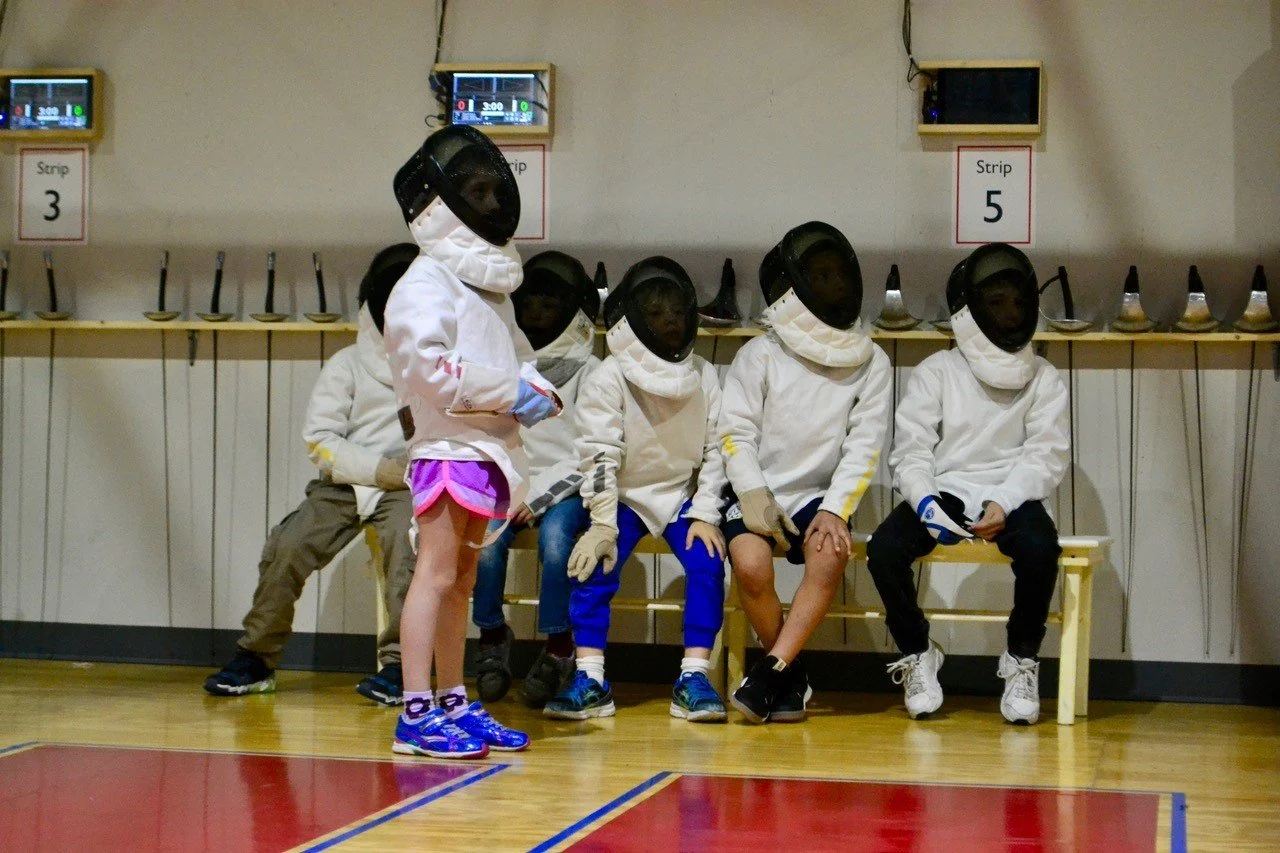 A young group of fencers waits on a bench for their chance to fence