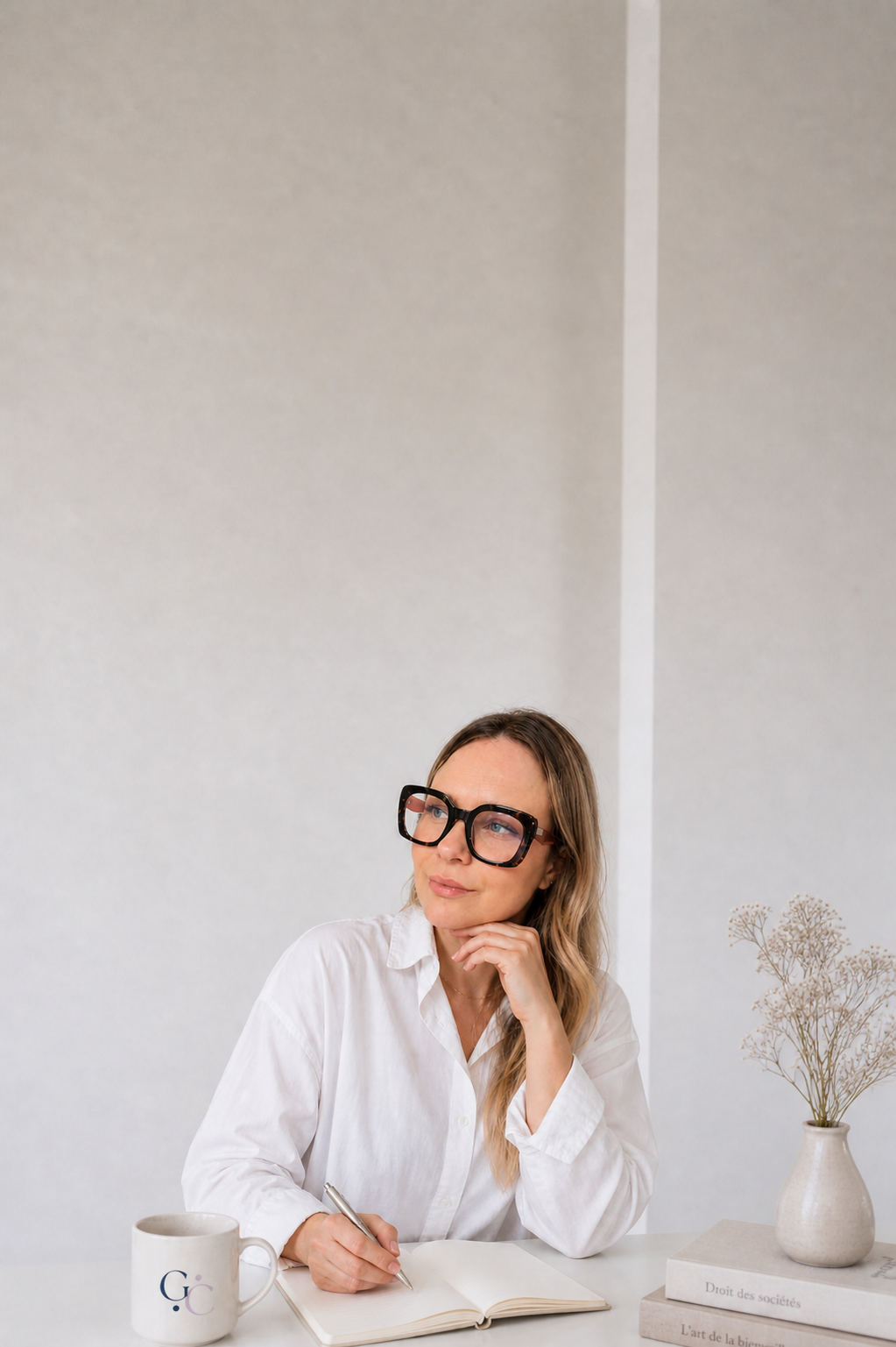 Une femme assise à une table blanche, portant des lunettes épaisses, en train d'écrire dans un carnet avec une tasse et une pile de livres à côté, dans un décor minimaliste avec une vase contenant des fleurs séchées.