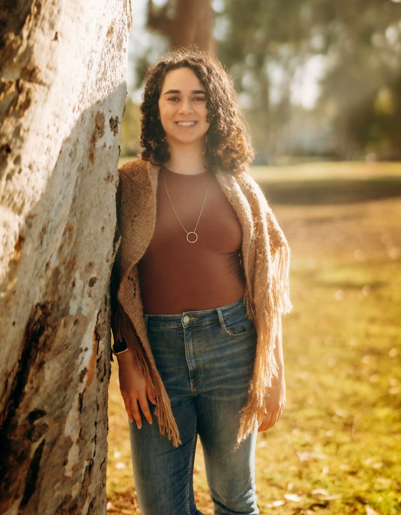 A young woman with curly hair smiling outdoors, leaning against a tree with sunlight in the background, wearing a brown shirt, a beige cardigan, and blue jeans.