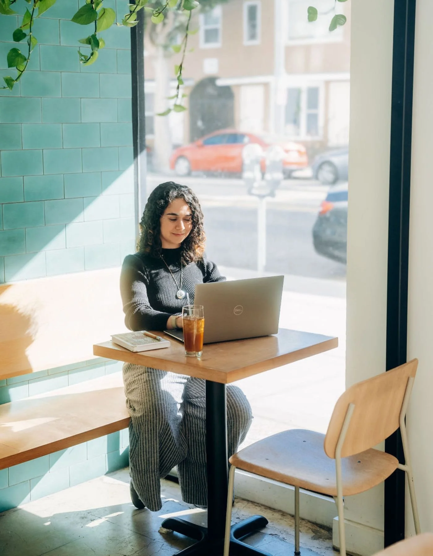 An online BIPOC therapist in CA sitting at a wooden table in a cafe with a laptop, a glass of iced drink, and a notebook. She is in natural light from large windows, with green tiled wall and hanging plant behind her, and city street visible outside.