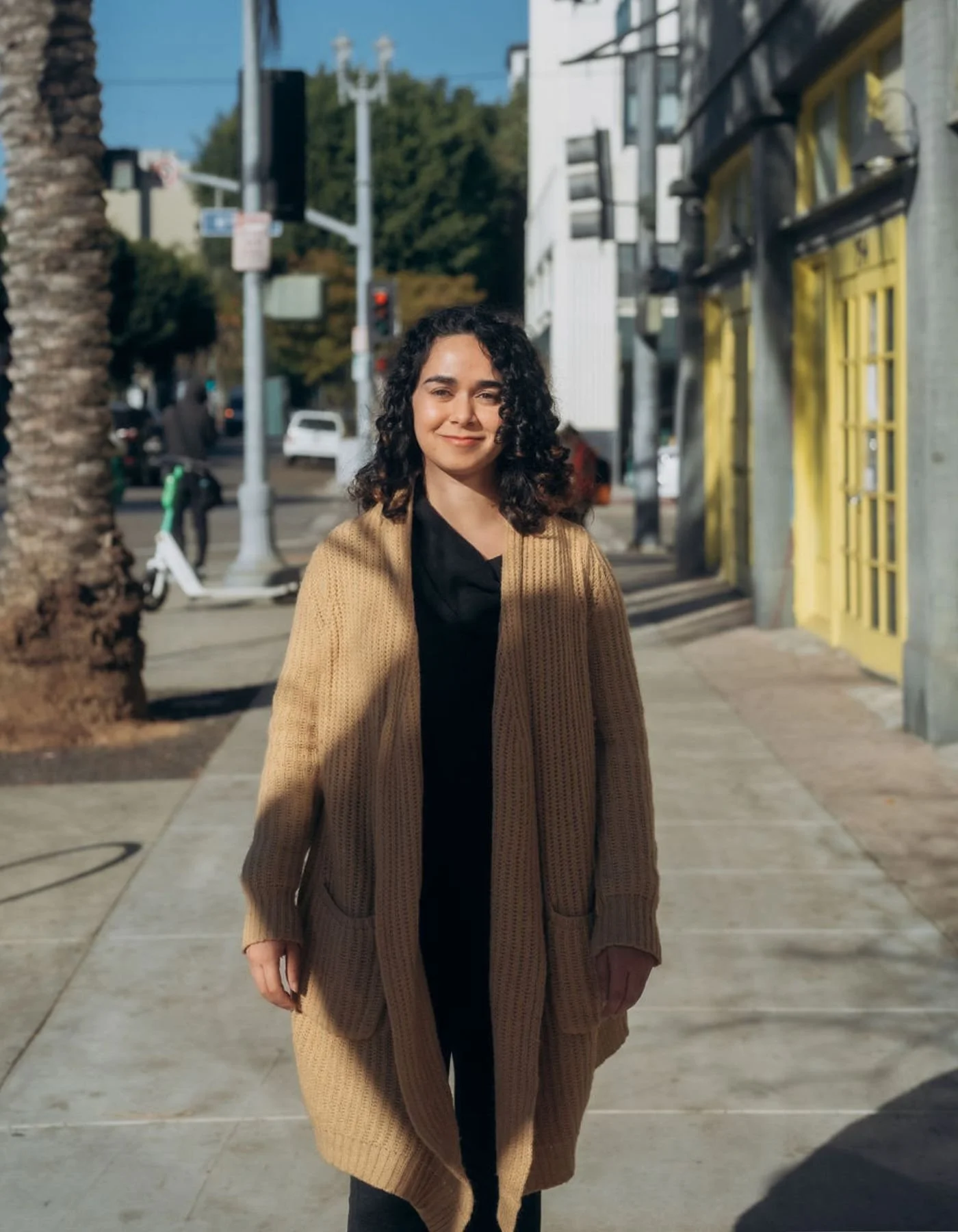 A young woman with dark curly hair wearing a tan knit coat over a black shirt standing on a city sidewalk with buildings, trees, and a parked bike in the background on a sunny day.