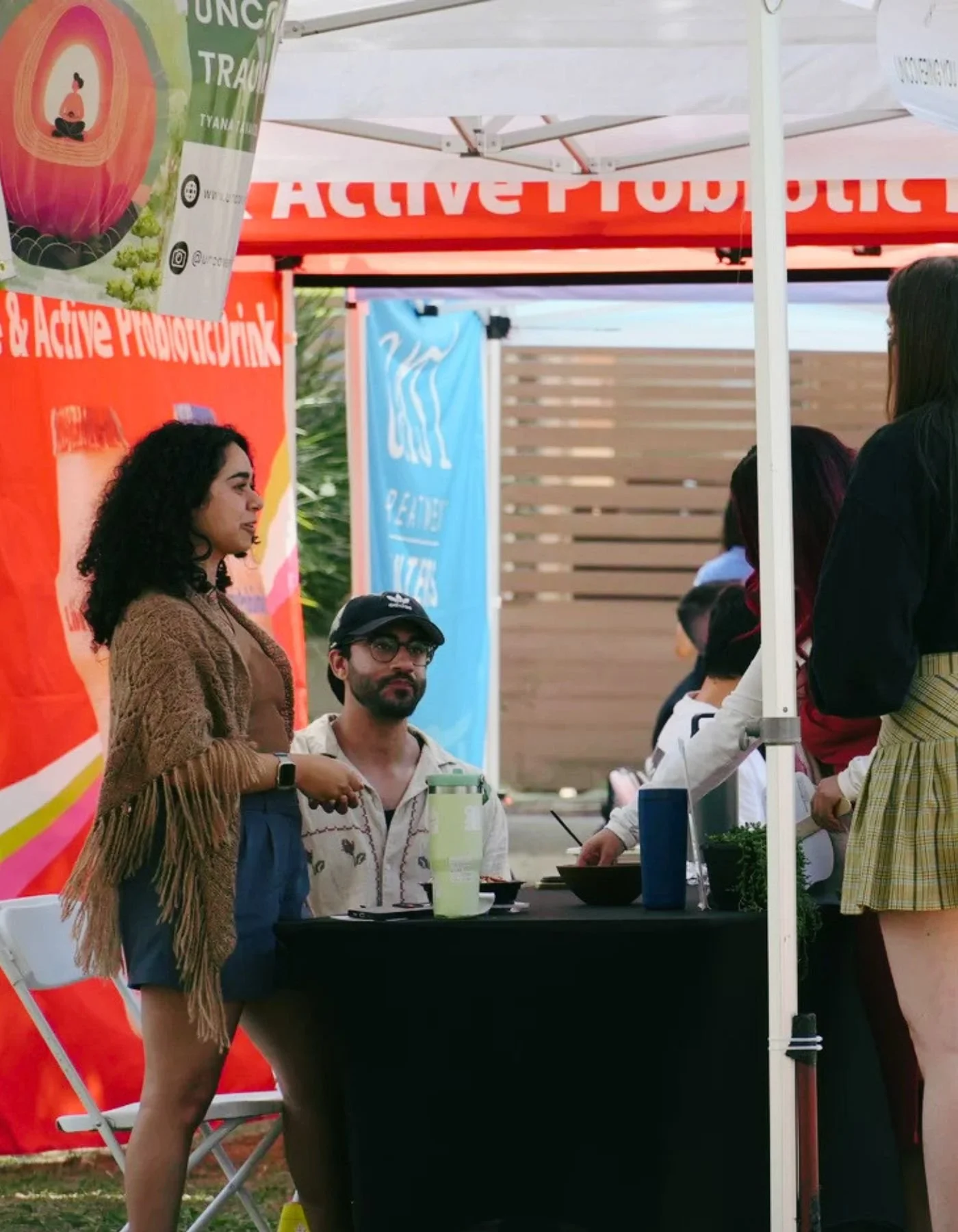 People standing and sitting at an outdoor booth under a white canopy tent with banners and red signage, engaging in conversation and viewing items on the table. Mental health expert and speaker for trauma, anxiety, neurodivergence (autism, ADHD).