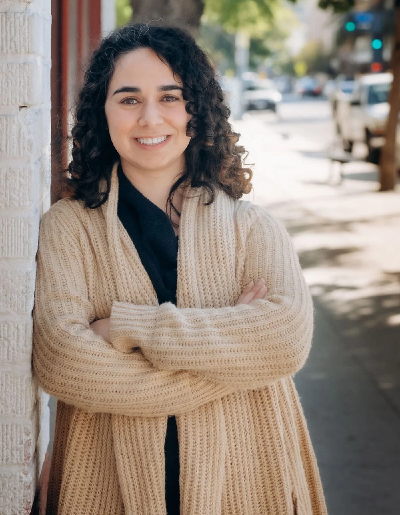 An anxiety therapist who does online self esteem therapy in California and Floriday with dark curly hair and a warm smile standing outdoors on a city street, leaning against a brick wall, wearing a beige knitted cardigan and a black top.