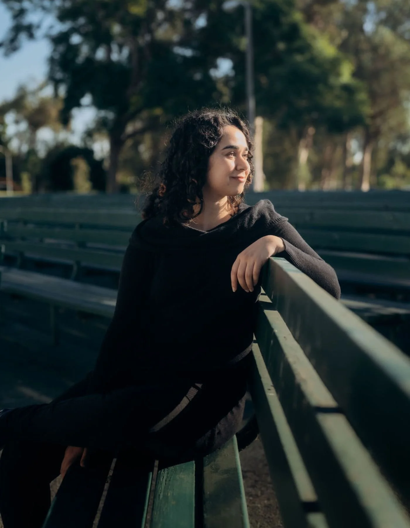 An online people pleaser therapist serving West Los Angeles and across California and Florida with curly dark hair leaning on a green park bench, looking to her right, with trees and sunlight in the background.