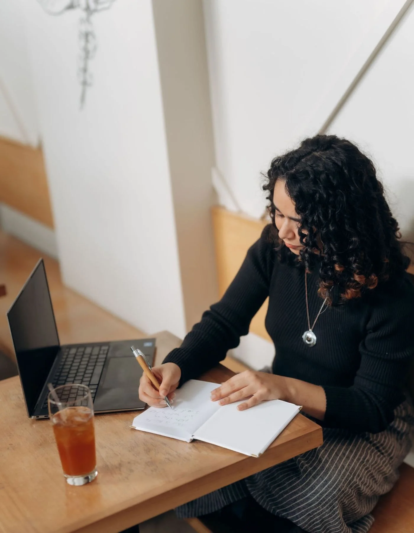 An online anxiety therapist in CA & FL with curly black hair writing in a notebook at a wooden table with a laptop, a glass of iced tea, and a pen. She is wearing a black sweater and a silver necklace, and appears to be in an indoor setting.