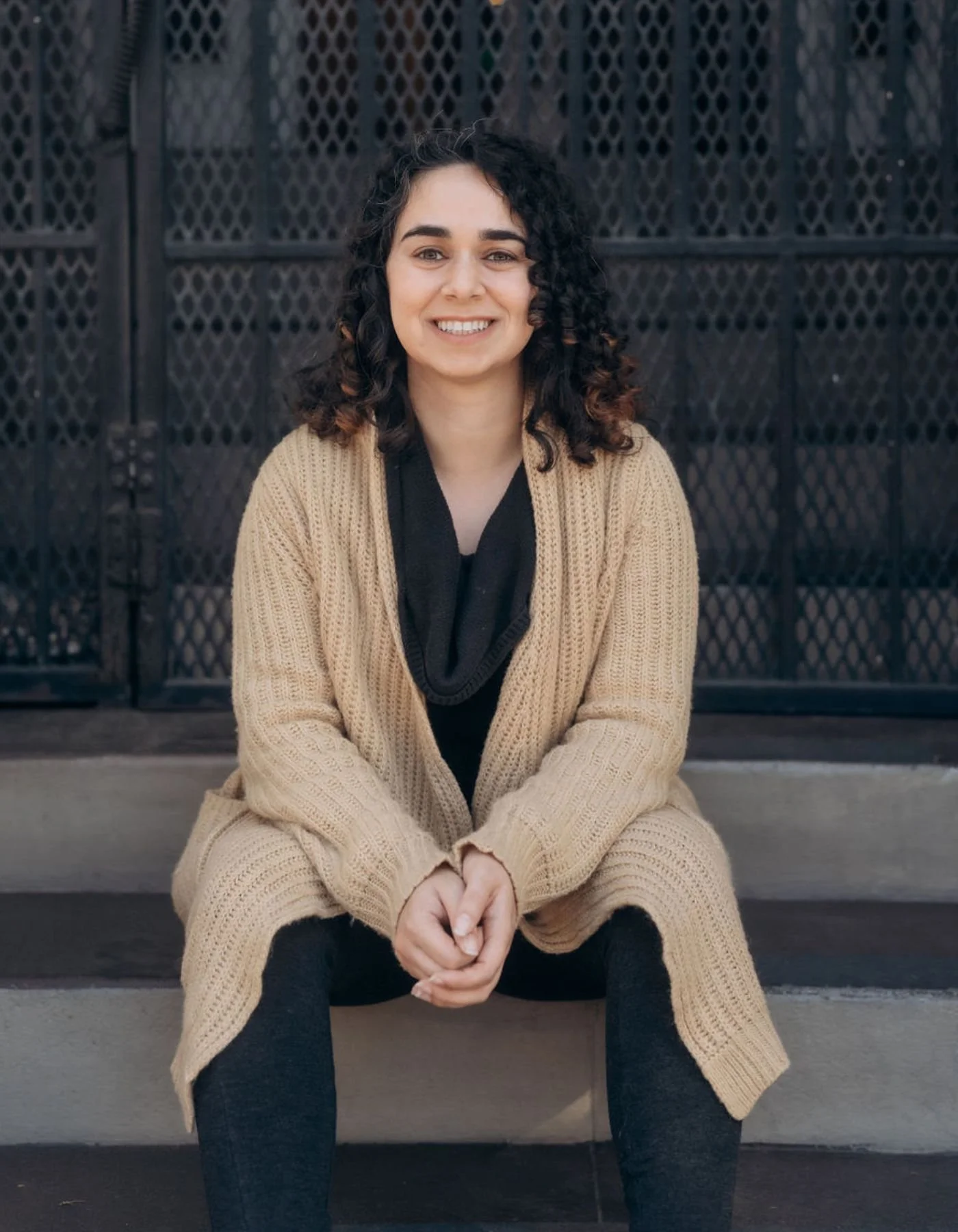 A BIPOC, neurodivergent affirming therapist in West Los Angeles, CA & FL who specializes in anxiety, people pleasing, self esteem, and HSP sitting on stairs in front of a black metal gate, smiling, wearing a tan cardigan and black pants with socks.