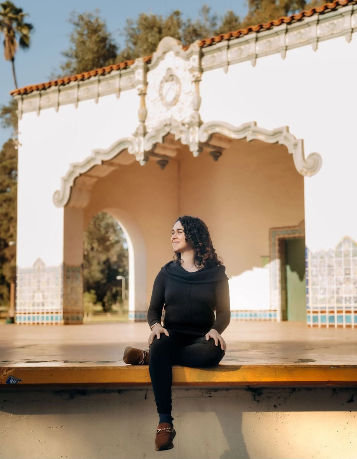 A woman with curly hair wearing a black shirt, black pants, and brown shoes sitting cross-legged on a stage platform, smiling and looking to her right. Behind her is an ornate archway with decorative detailing, part of a historical building with Spanish-style architecture. Trees and a clear sky are in the background.