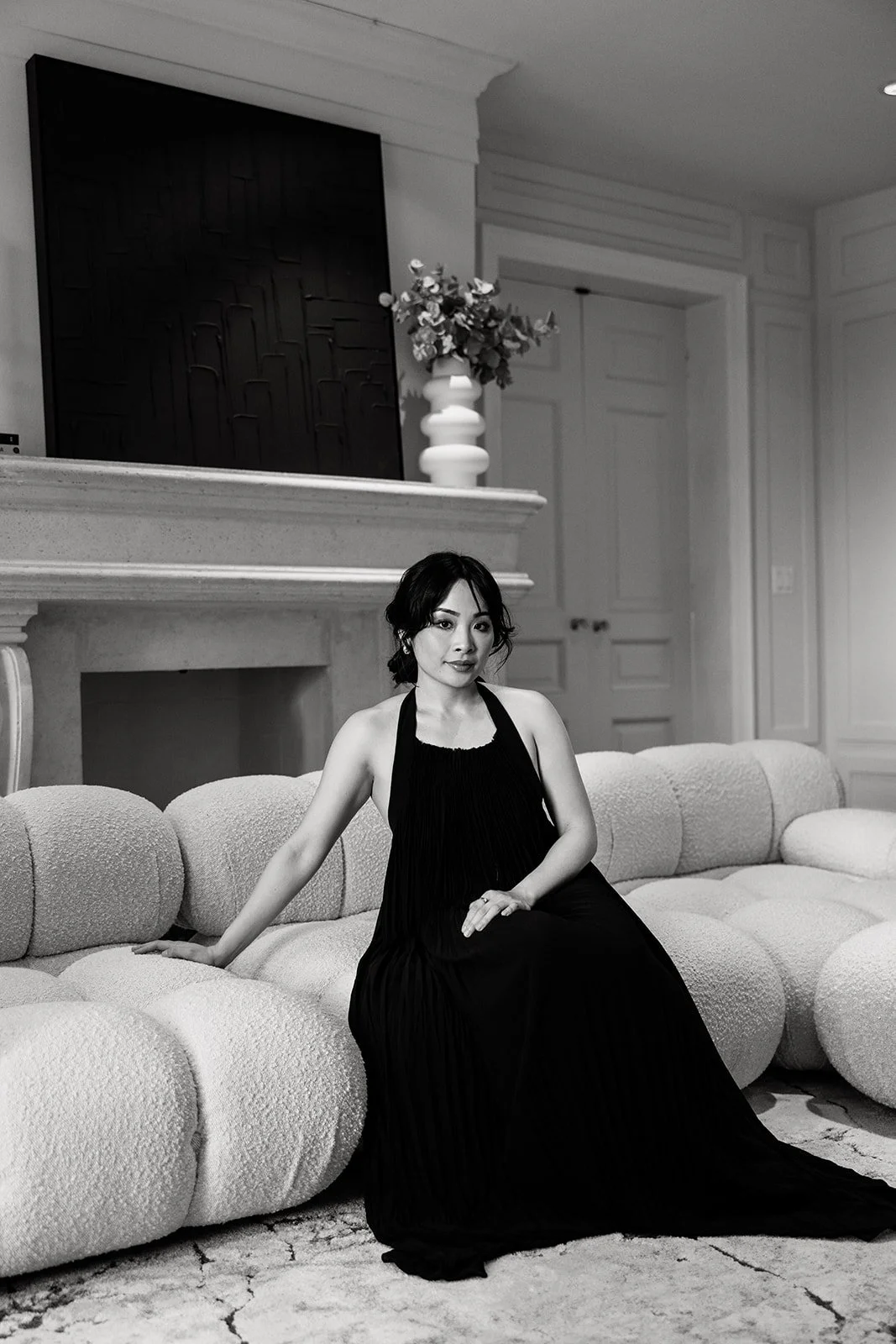 A woman in a black dress sitting on a textured, rounded couch in a well-decorated living room, black-and-white photo.