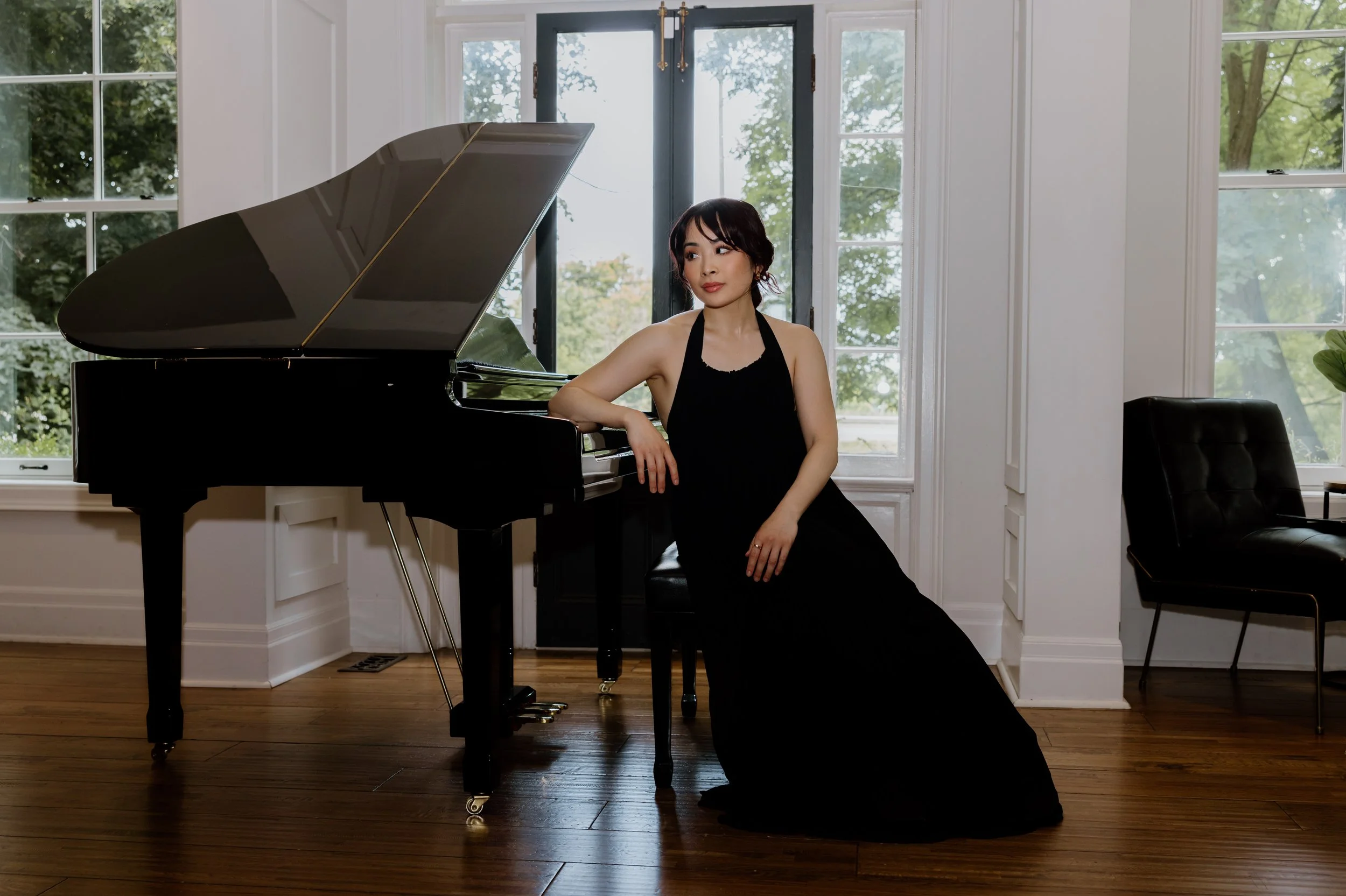 A woman with dark hair wearing a black dress sitting next to a grand piano in a bright room with large windows showing green trees outside.
