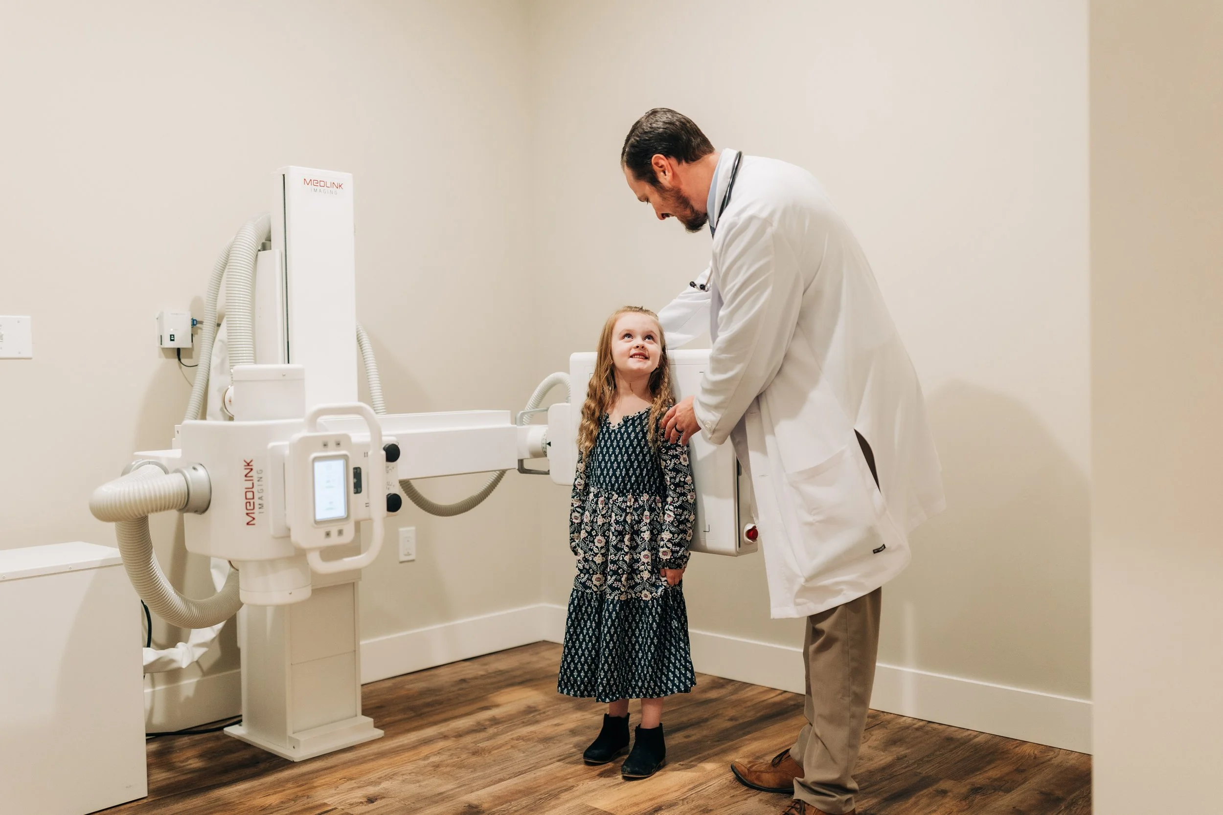 A doctor examining a young girl in a medical imaging room, with medical equipment and a beige wall in the background.