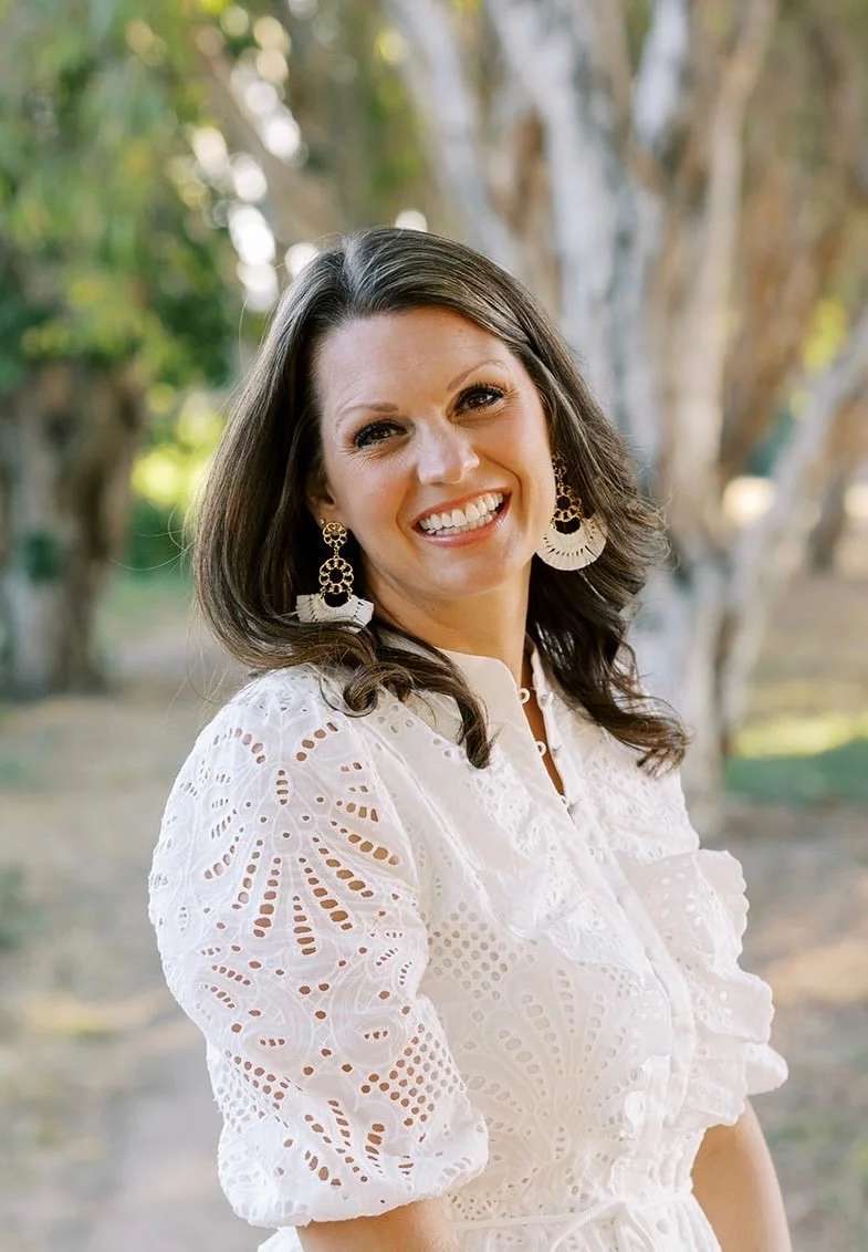 A woman smiling outdoors, wearing a white eyelet dress and large earrings, with trees in the background.